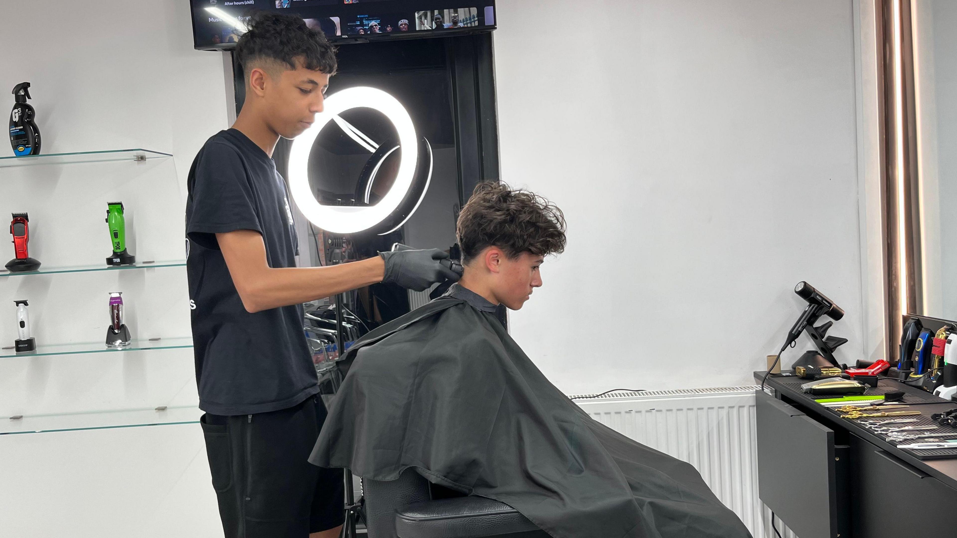 A boy is sitting on a barber's chair with a black gown on. Another boy is cutting his hair and is wearing black gloves.