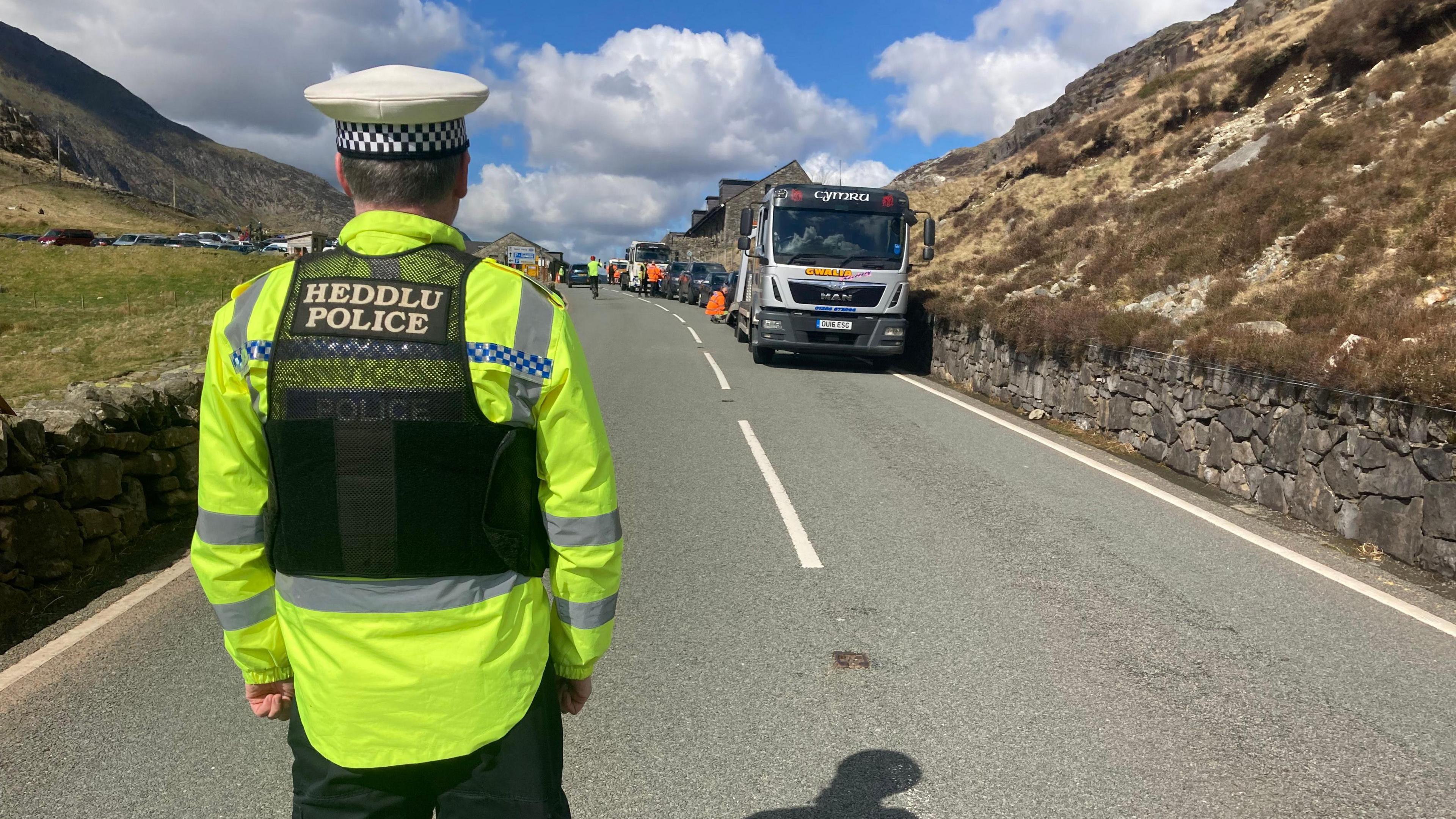 Police officer with his back to camera watches as a recovery vehicle is loaded with a car being towed away for illegally parking at Pen y Pass near Llanberis