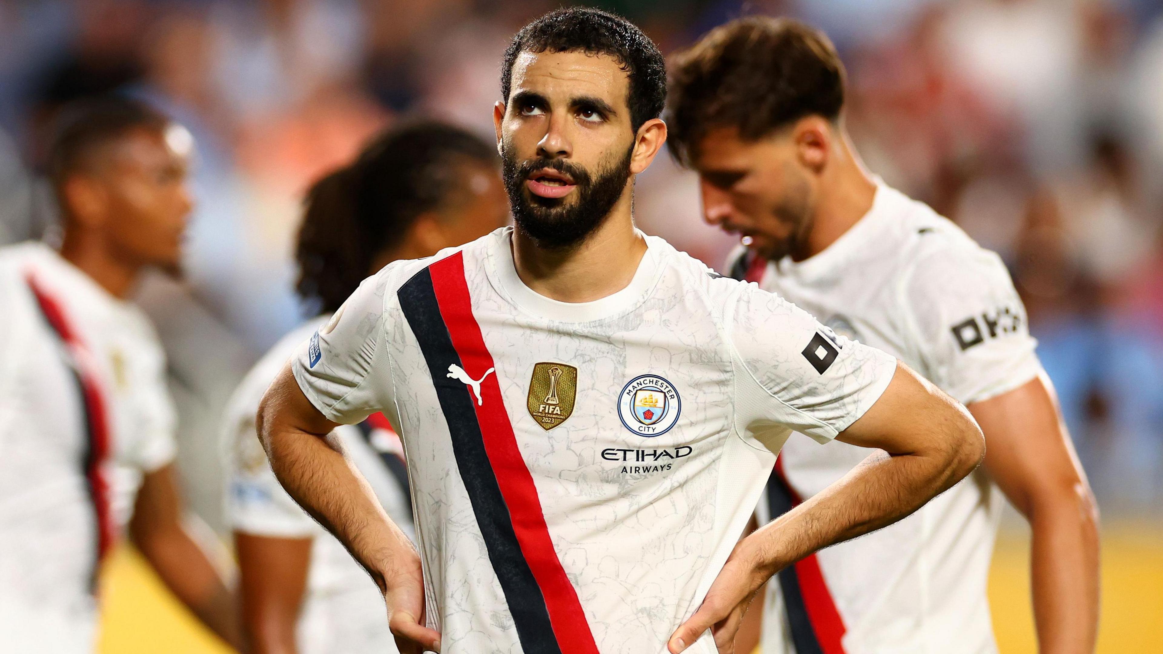 Rayan Ait-Nouri, yllään Manchester City Cream -paita, jossa musta ja punainen diagonaalivyönauha seisoo hänen lantiossaan ja etsii jalkapallo-ottelua, kolme näkyvää, mutta hämärtää joukkuetoveria takanaan