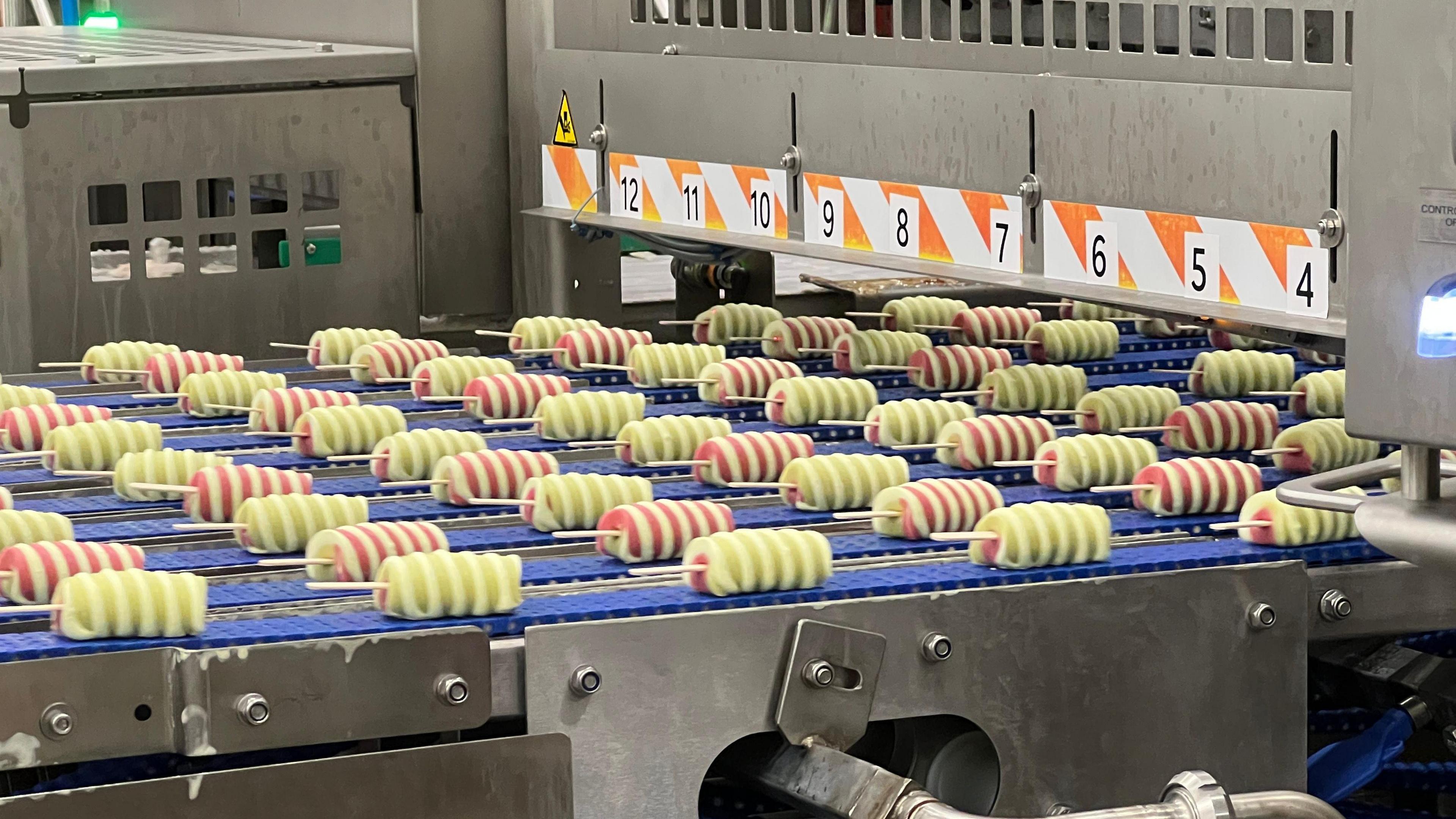 Twister ice cream on sticks, which are stripped in colour, travelling down a conveyor belt in a factory. 