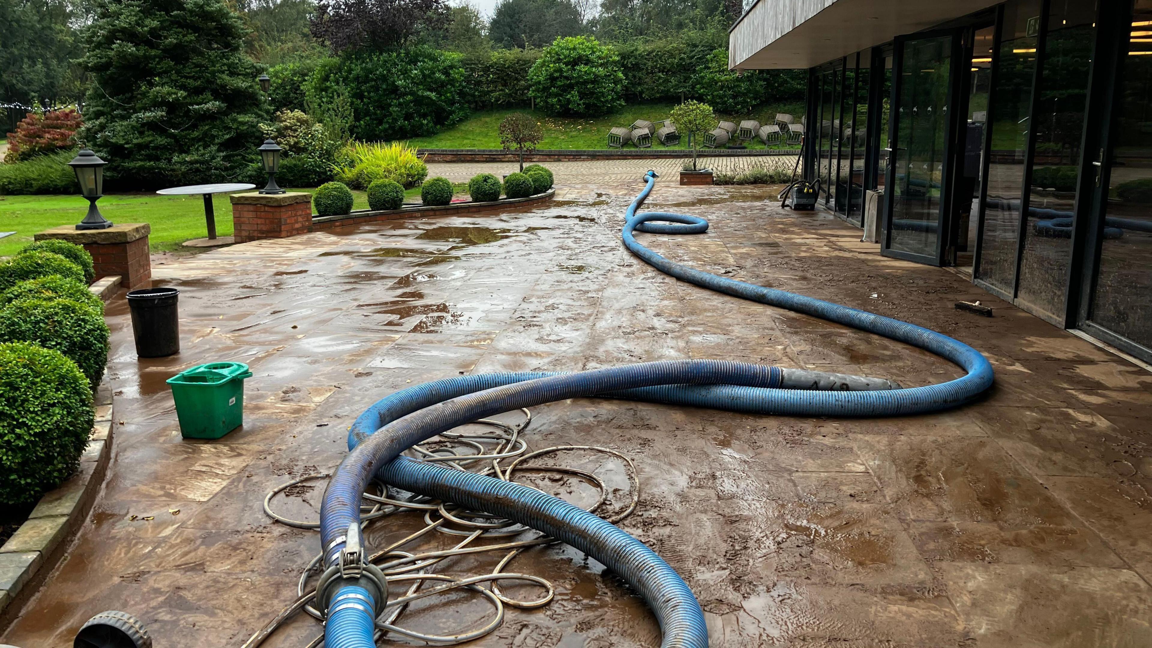 A long, muddy blue pipe on a patio between a building with glass windows and a garden.