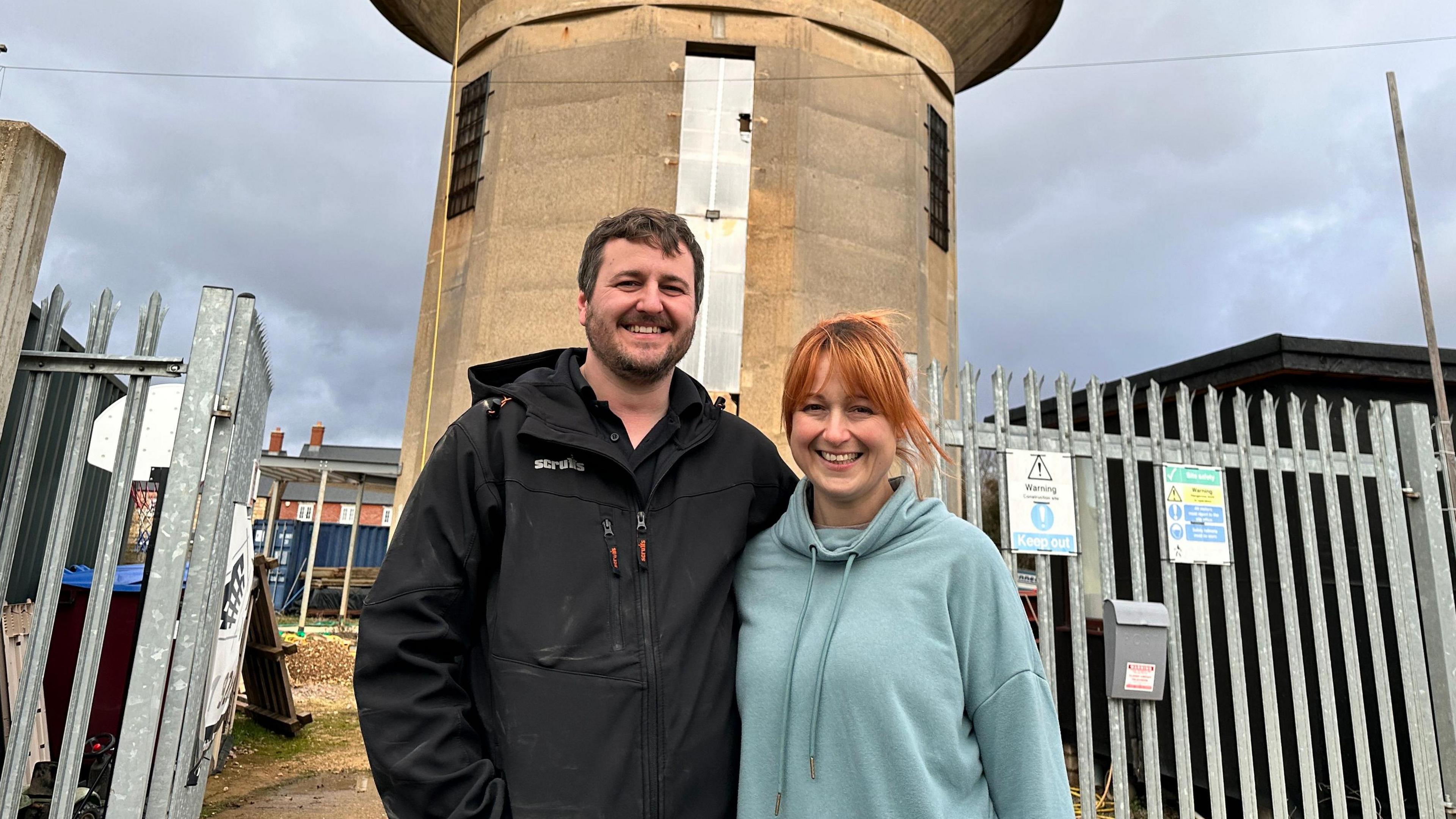 Adam, in a black jacket, stands next to Tassy, who has orange hair and is wearing a blue hoodie. They are standing in front of a gate in a metal fence. The stone water tower is behind them. 