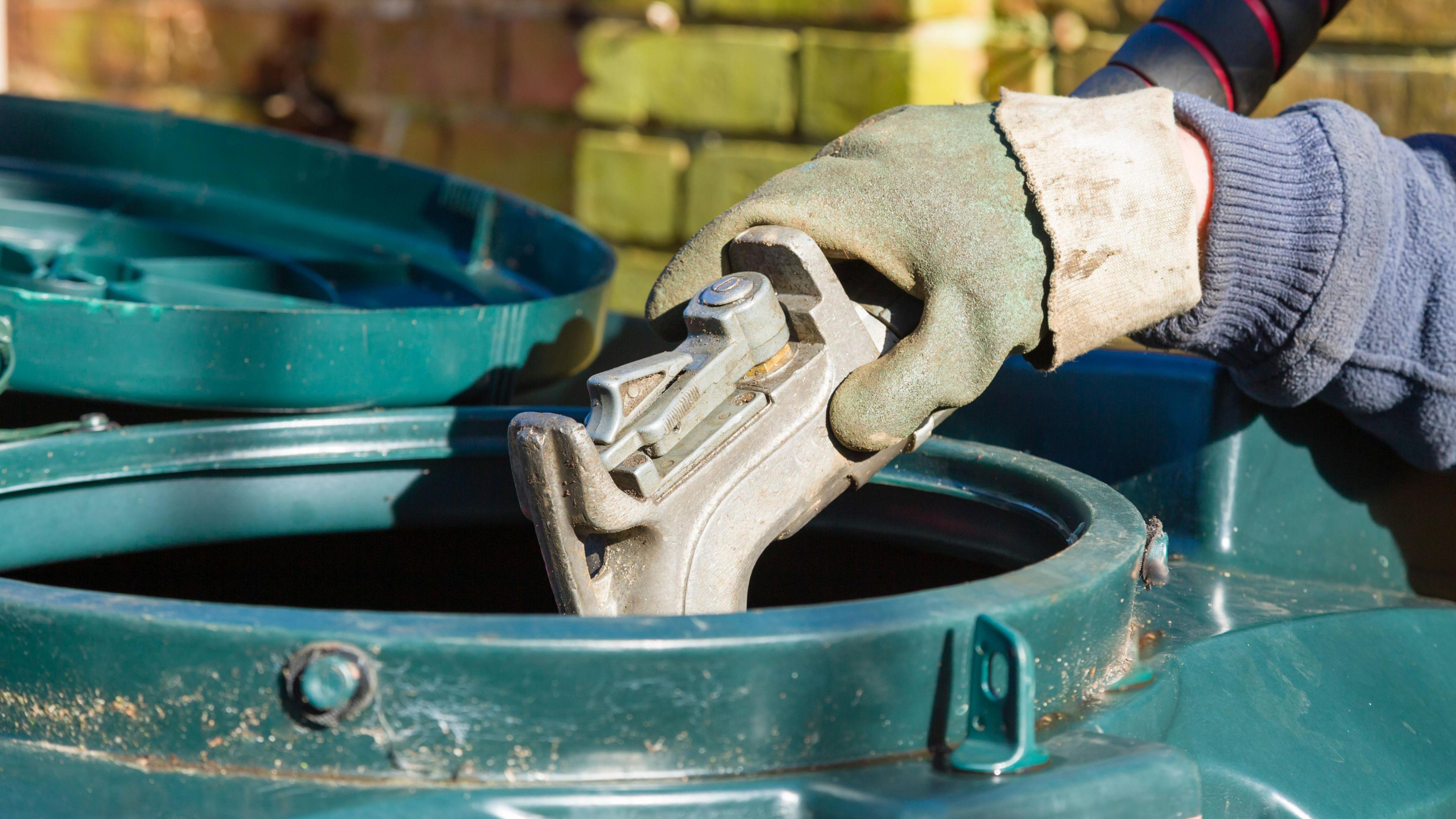 A gloved hand holds a metal pump in the opening of a green oil tank