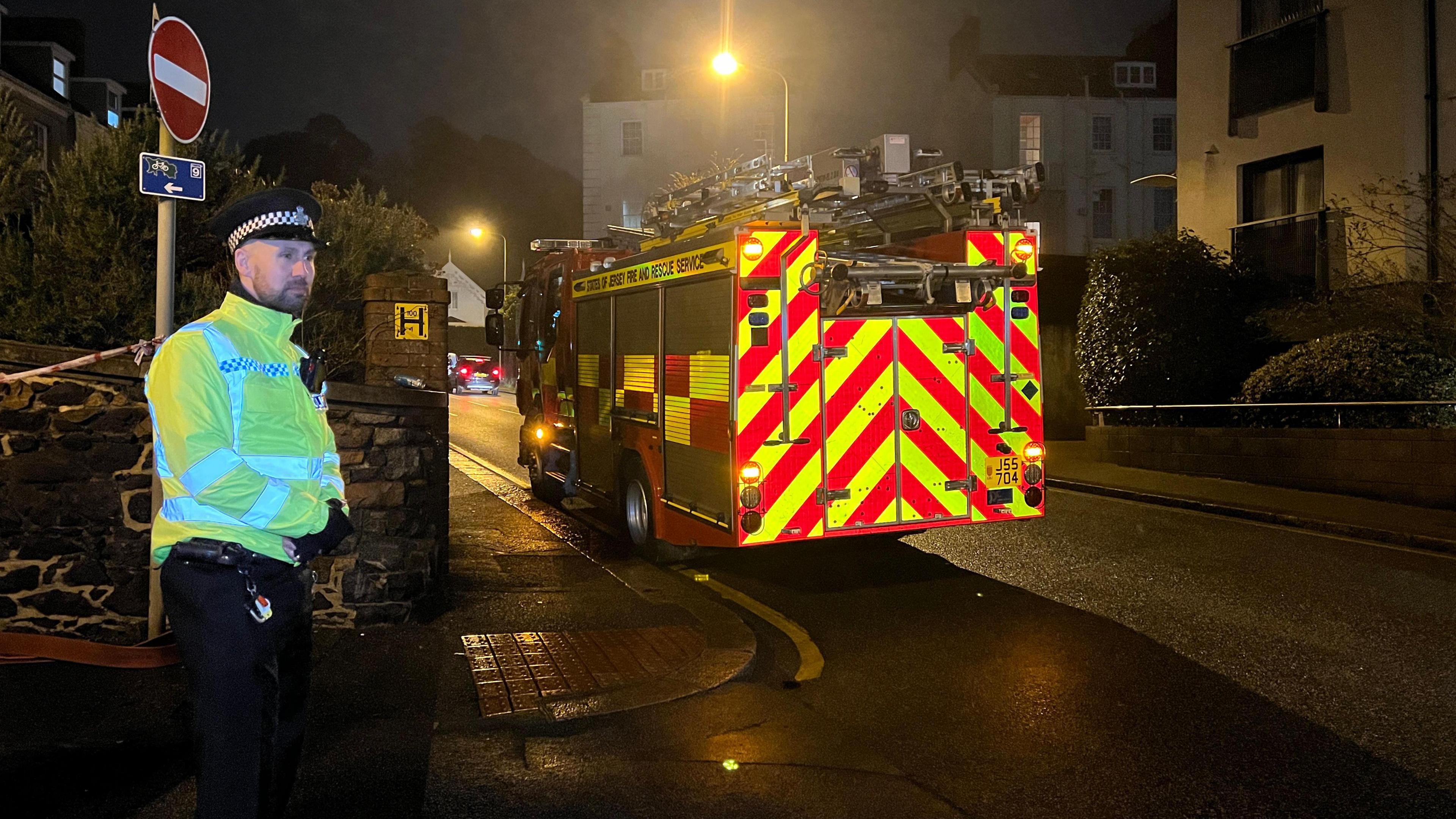 A policeman in high-viz stands to the left of the image, while a fire engine can be seen to the right. It is dark.