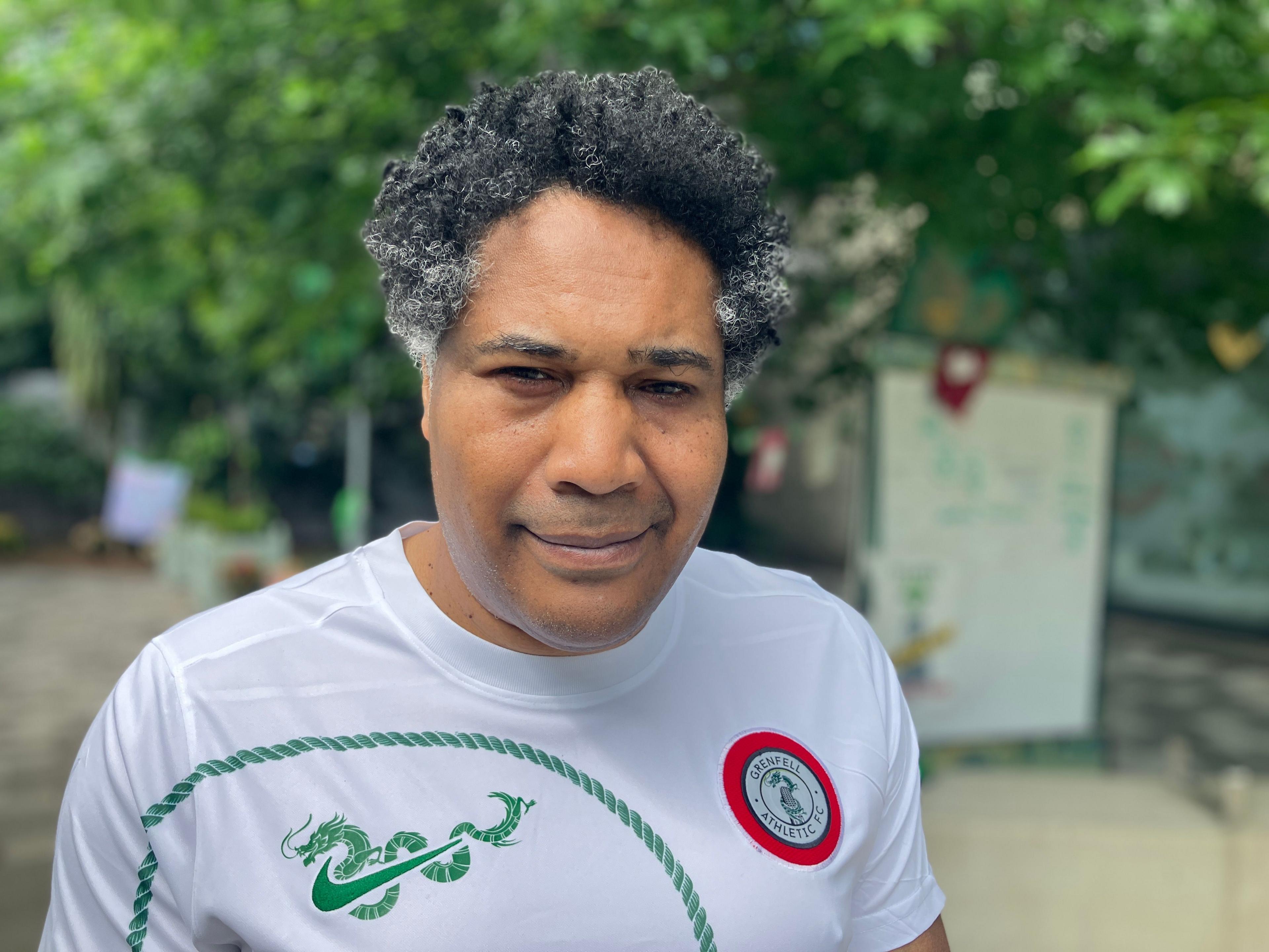 A man with curly black-grey hair smiles at the camera. He is wearing a white T-shirt with a green dragon logo on it. In the background out-of-focus greenery can be seen