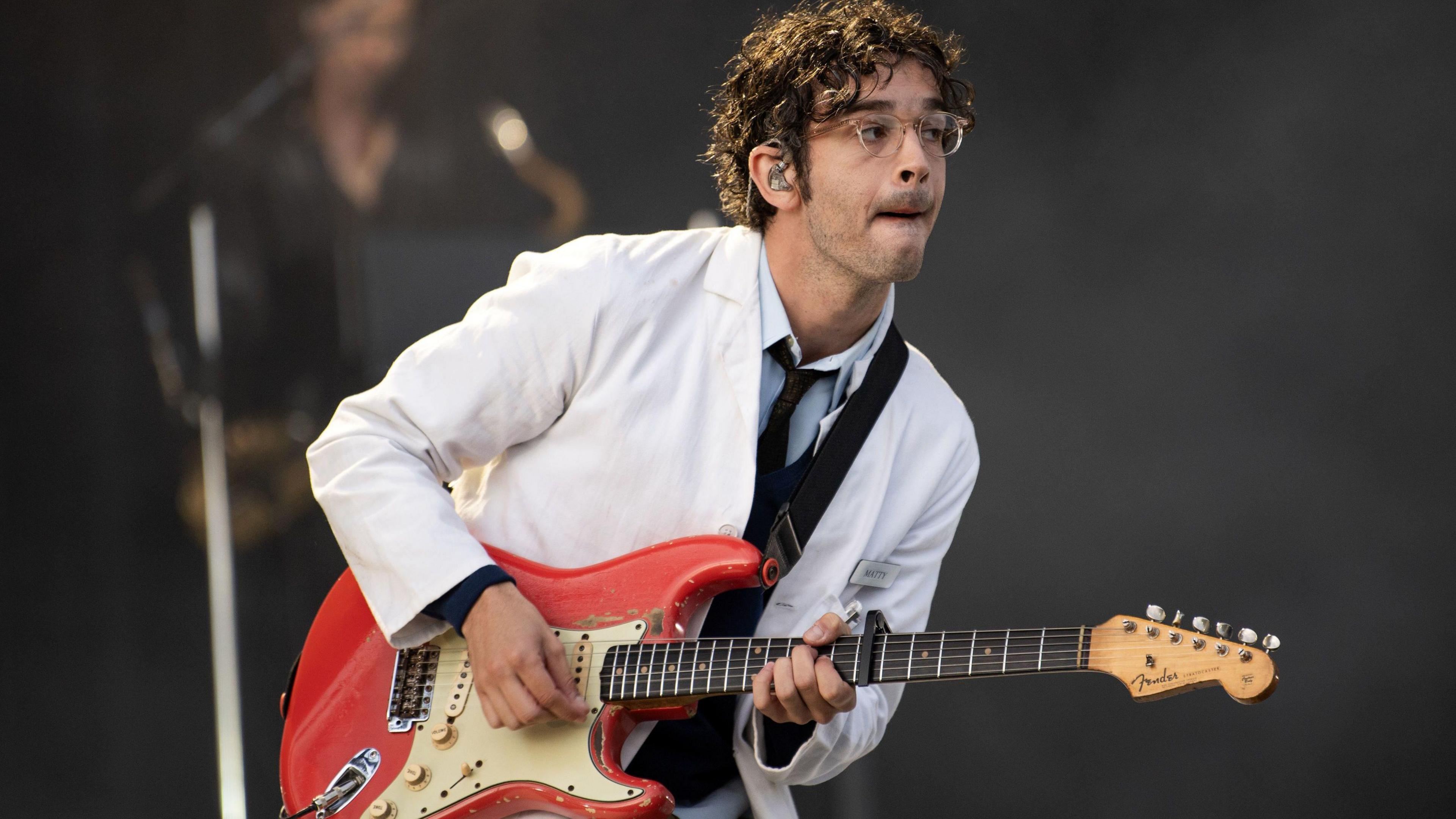 The 1975's Matty Healy, dressed in a white suit, pulls a face while playing guitar on stage at a festival in Scotland, 2024