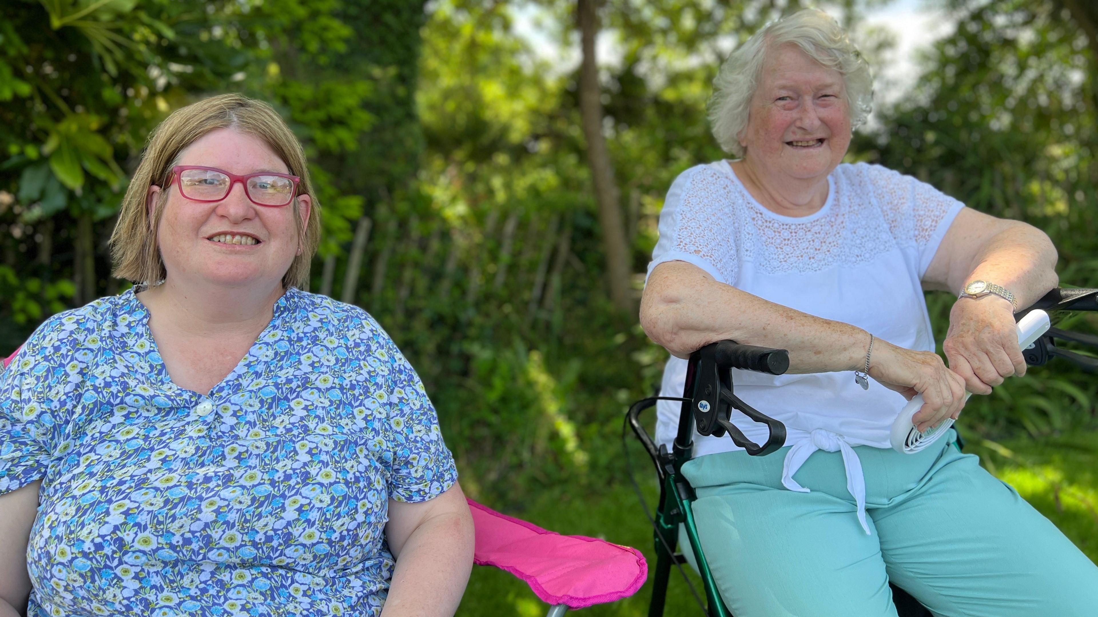 Two women sat facing the camera. The woman on the left is wearing a blue patterned top and has a bob and is wearing pink glasses. The woman on the right is wearing light blue trousers and a white top. Both are looking at the camera smiling. Behind then is green grass and trees.