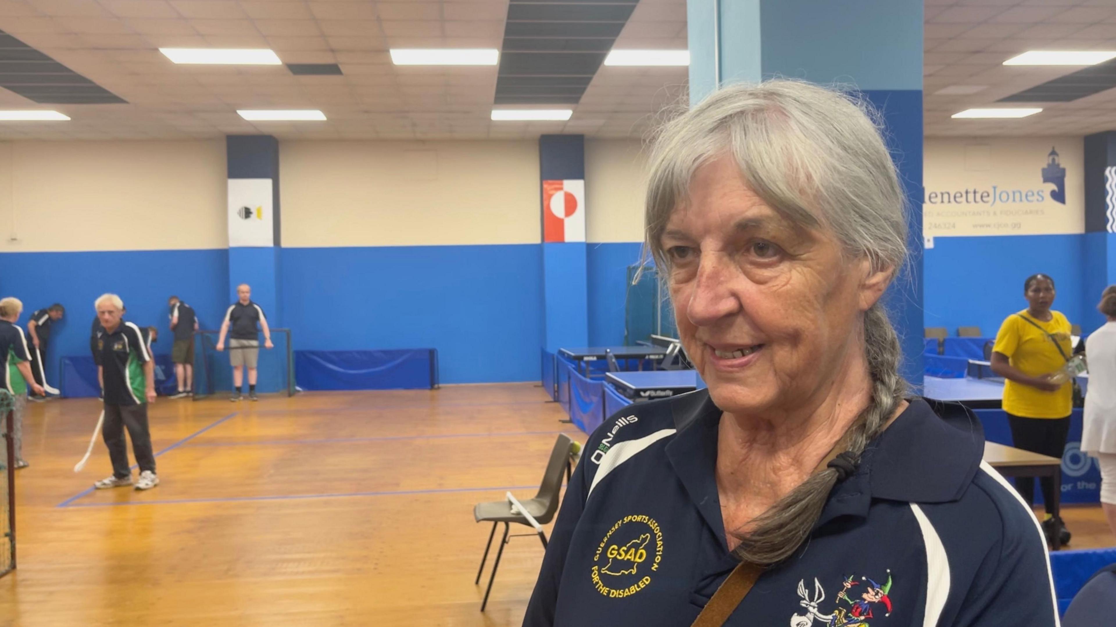 A woman in a blue tracksuit that says GSAD: Guernsey Sports Association for the Disabled. She is in a sports hall.