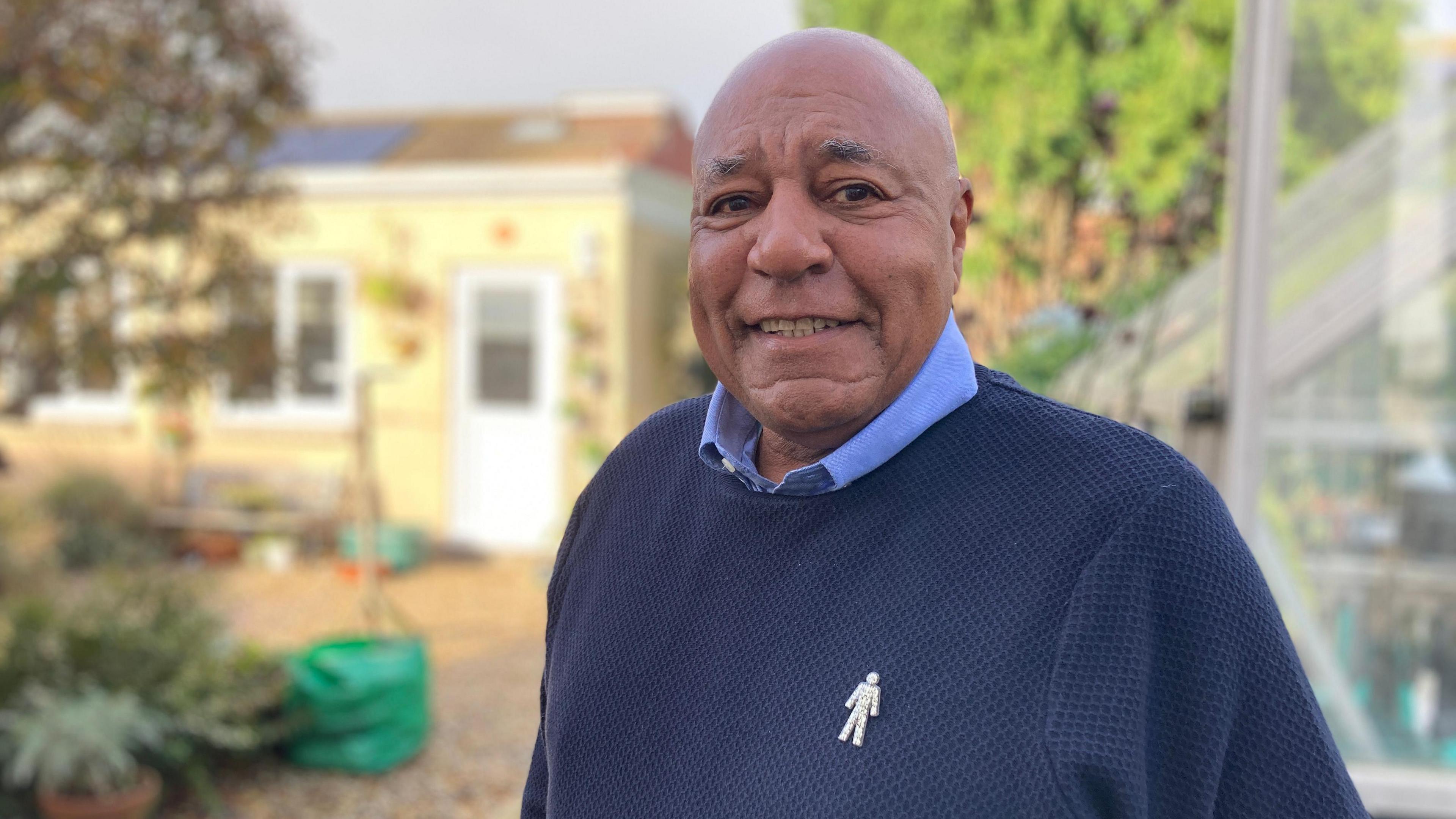 Julian Delaney, pictured in his garden in Vale of Glamorgan. He is wearing a dark blue jumper with a light blue shirt underneath and has a cancer badge in the outline of a man on the left hand side of his chest