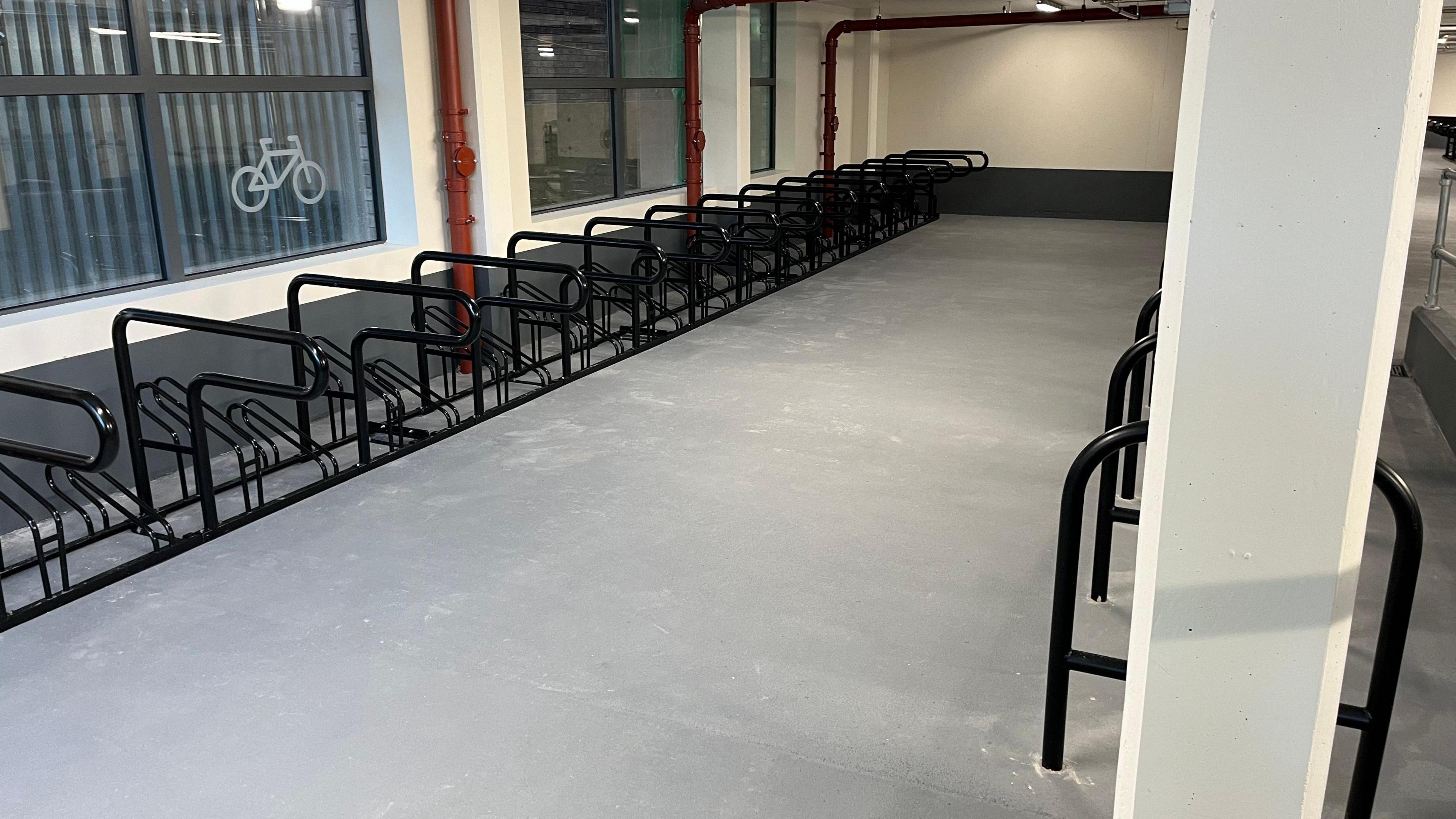 A grey room with white painted walls has black bike racks lined up next to each other. On one window is a white symbol for a bicycle.