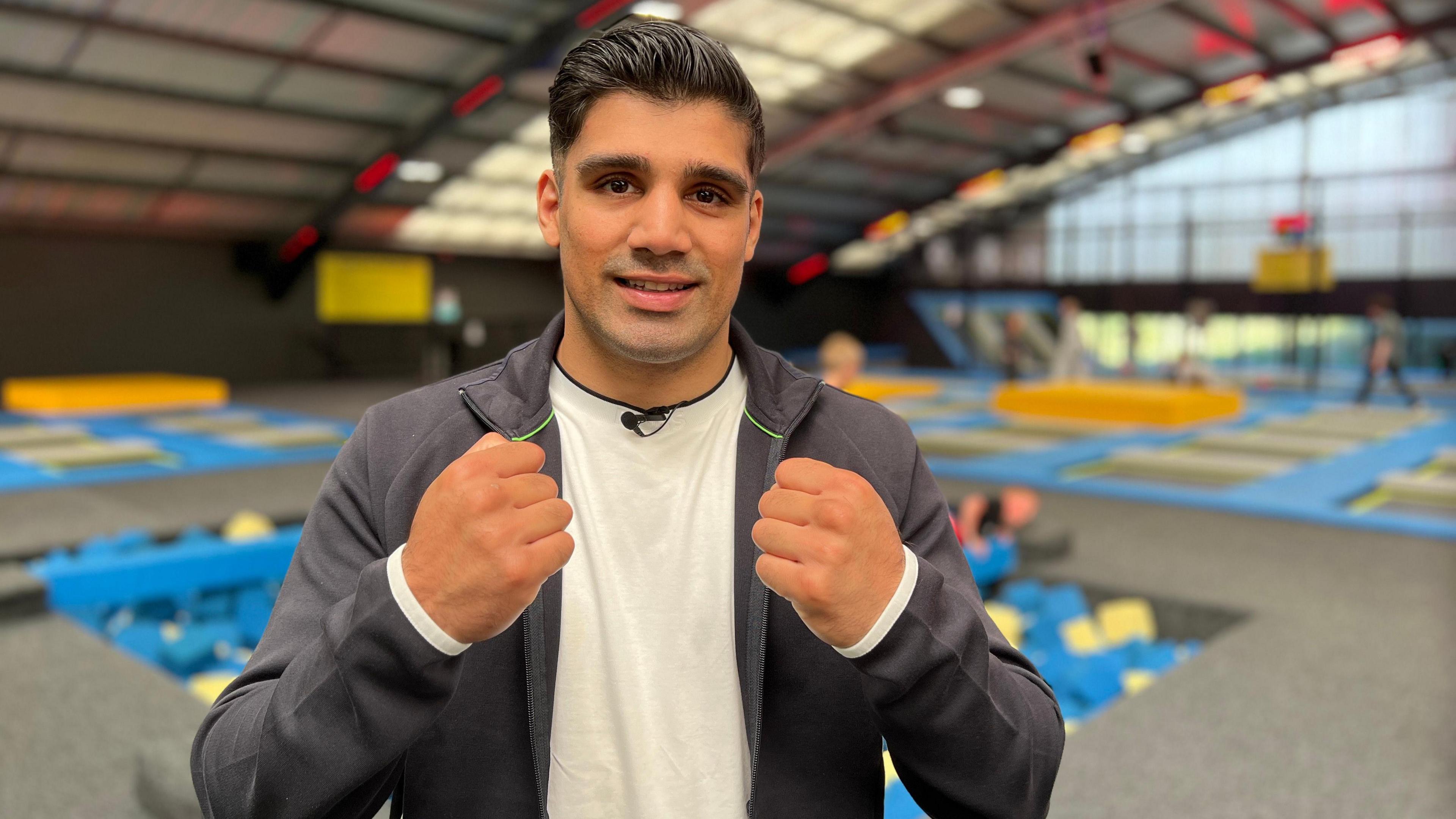 Muhammad looks at the camera with his hands up in the air in a boxing stance. He smiles as in the background there is a soft play area with trampolines where some children and their parents are playing.