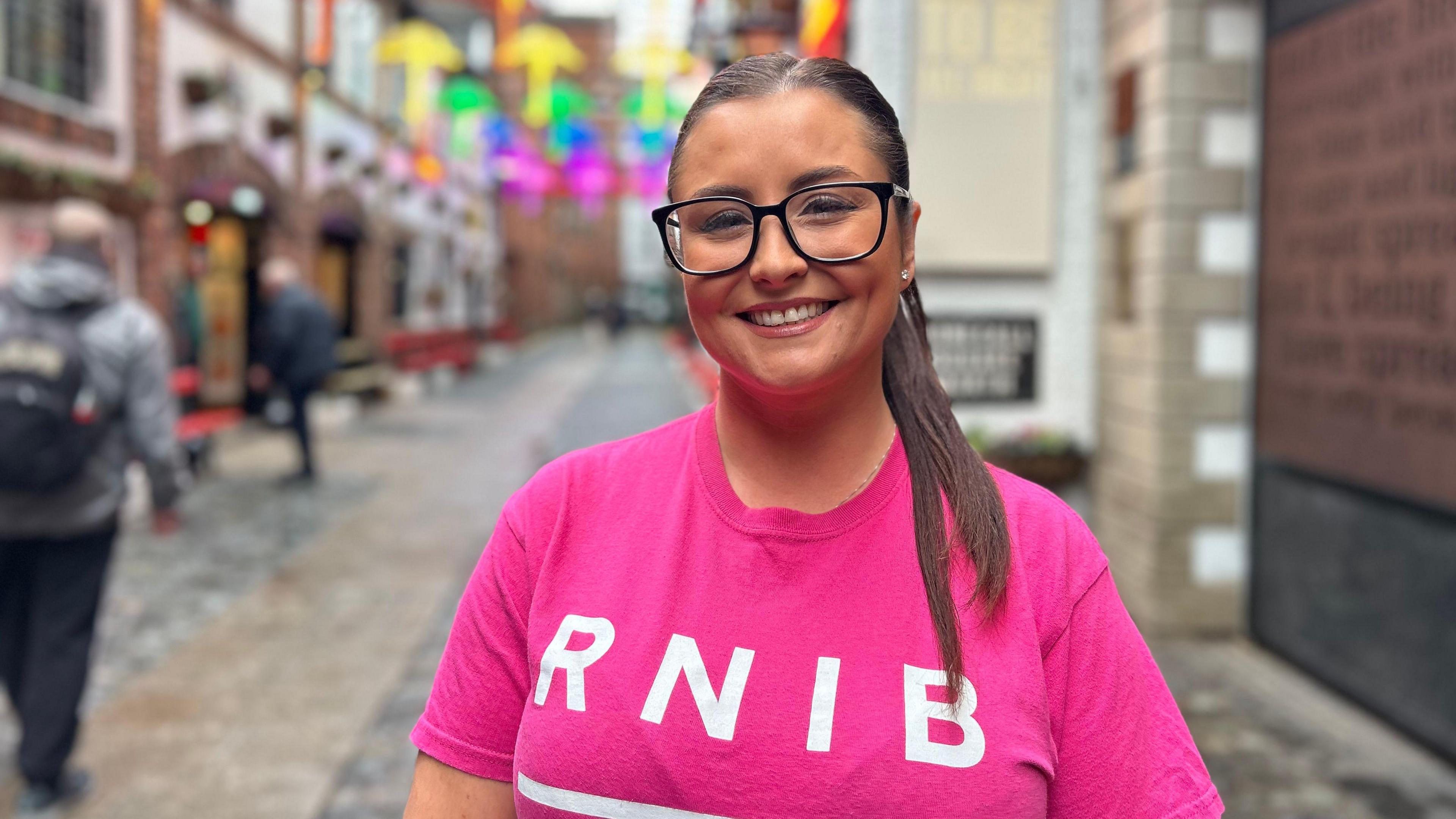 Laura Cummings pictured in Belfast's cathedral quarter wearing a pink RNIB shirt, she has long brown hair in a ponytail and glasses