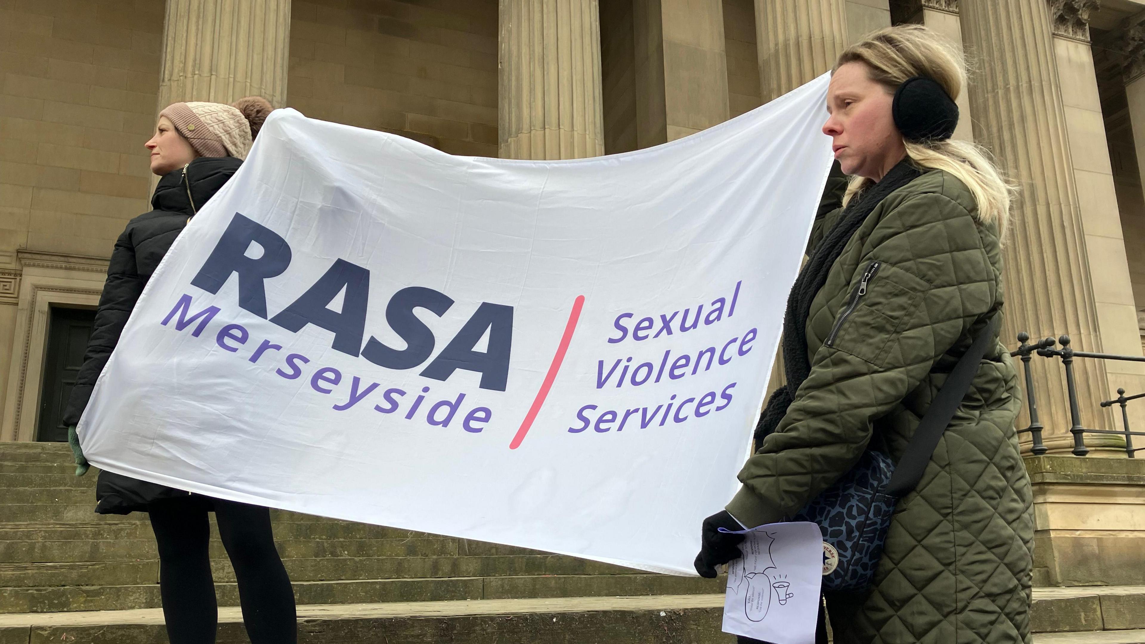 Two women hold up a white canvas with RASA Merseyside and sexual violence services written on it in purple lettering