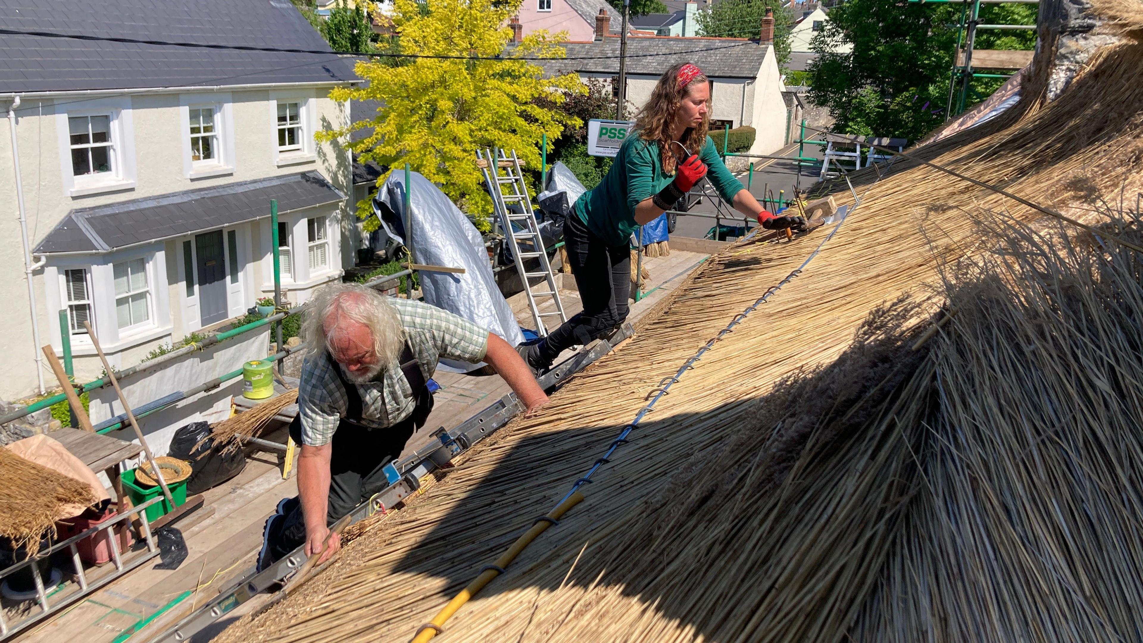 Alan Jones and Priya Pannochia on top of a thatched roof. They are both kneeling over and repairing the roof as the sun shines down on them. They both have tools in their hands.