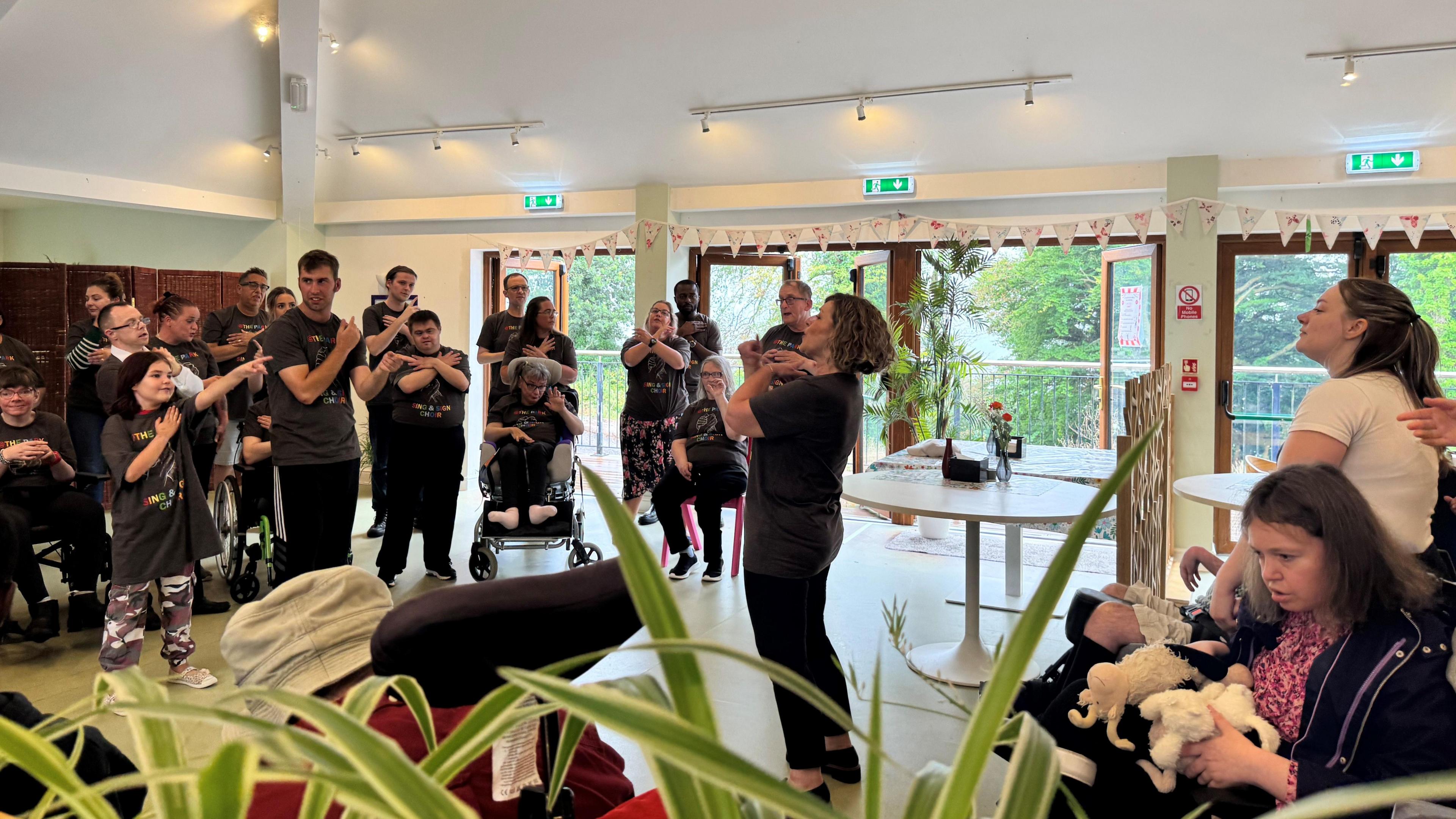 Group of people, singing and signing, as part of a choir to residents where they live. They are standing in a large room, wearing grey tee-shirts with the name of their choir on. The people who are watching them are either standing up or in wheelchairs.