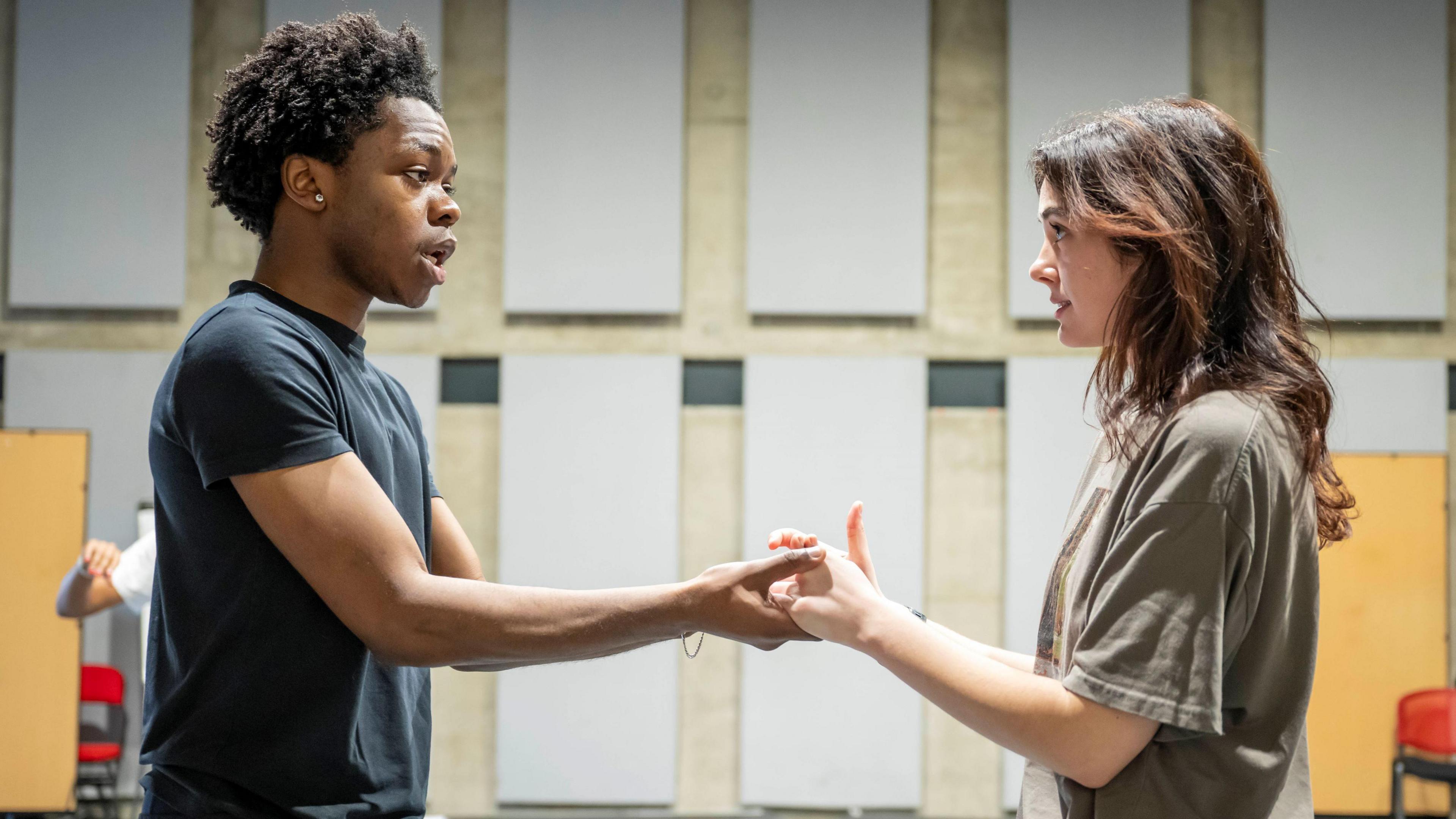 Two young actors, one male and one female, face each other and hold hands during rehearsals for Romeo and Juliet at Bristol Old Vic. They are wearing casual clothing