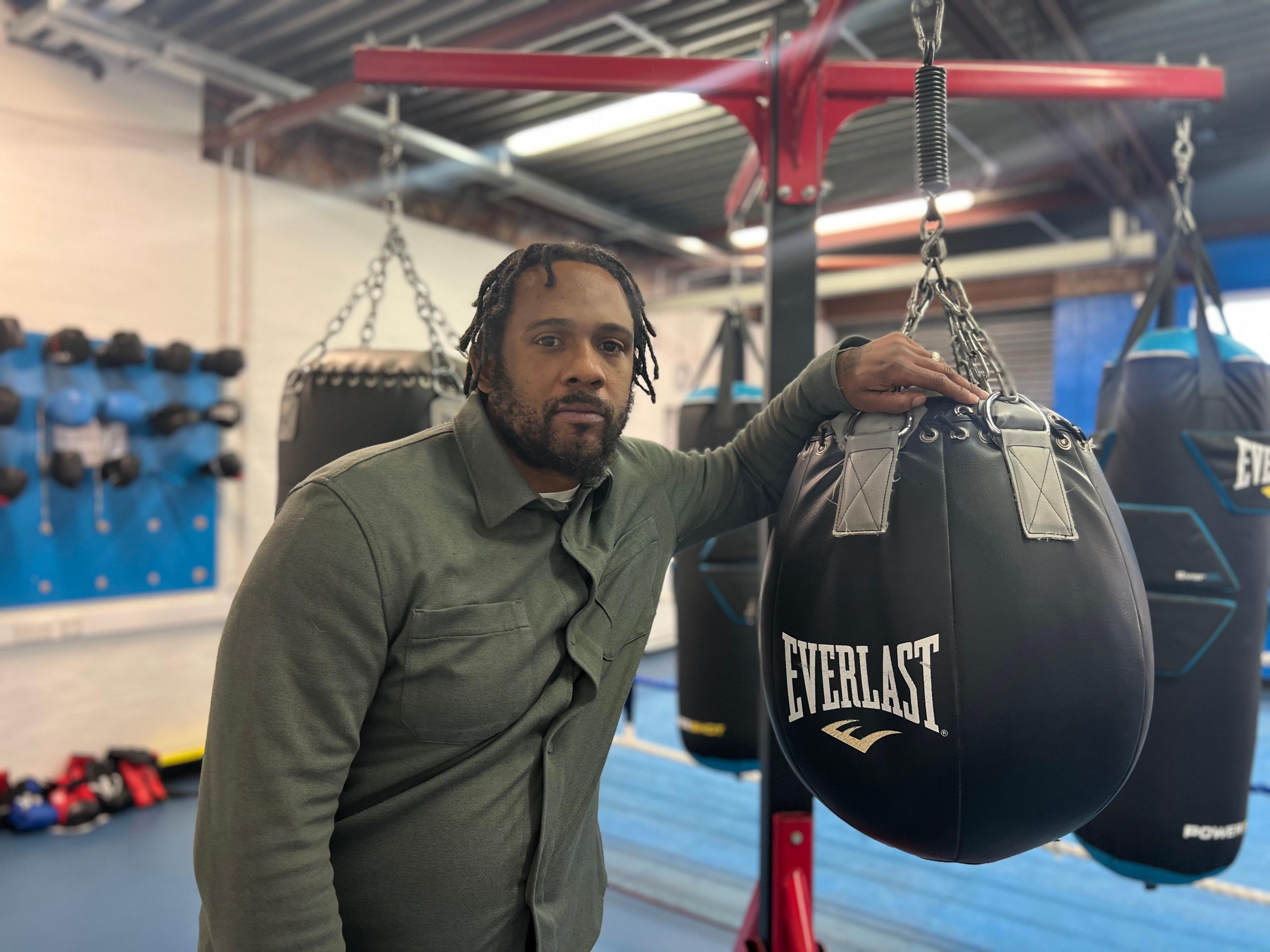 Dayton is standing next to a punching bag in a boxing gym. He has a green shirt on and wears his hair in braids. 