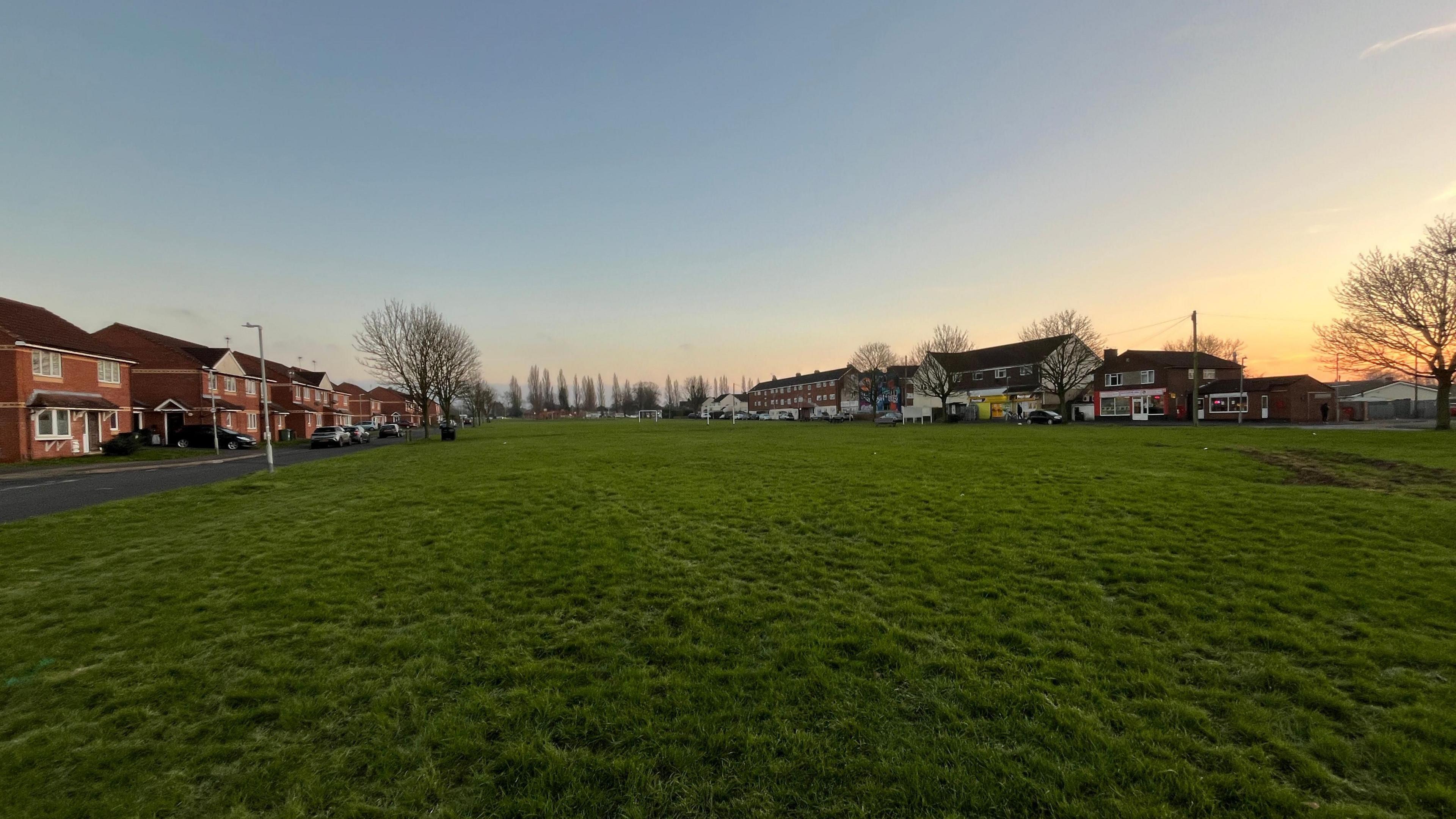 A large green space in an urban area during a clear winter sunset. There are houses and shops surrounding the green space, which is mostly just an empty field, with trees in the background