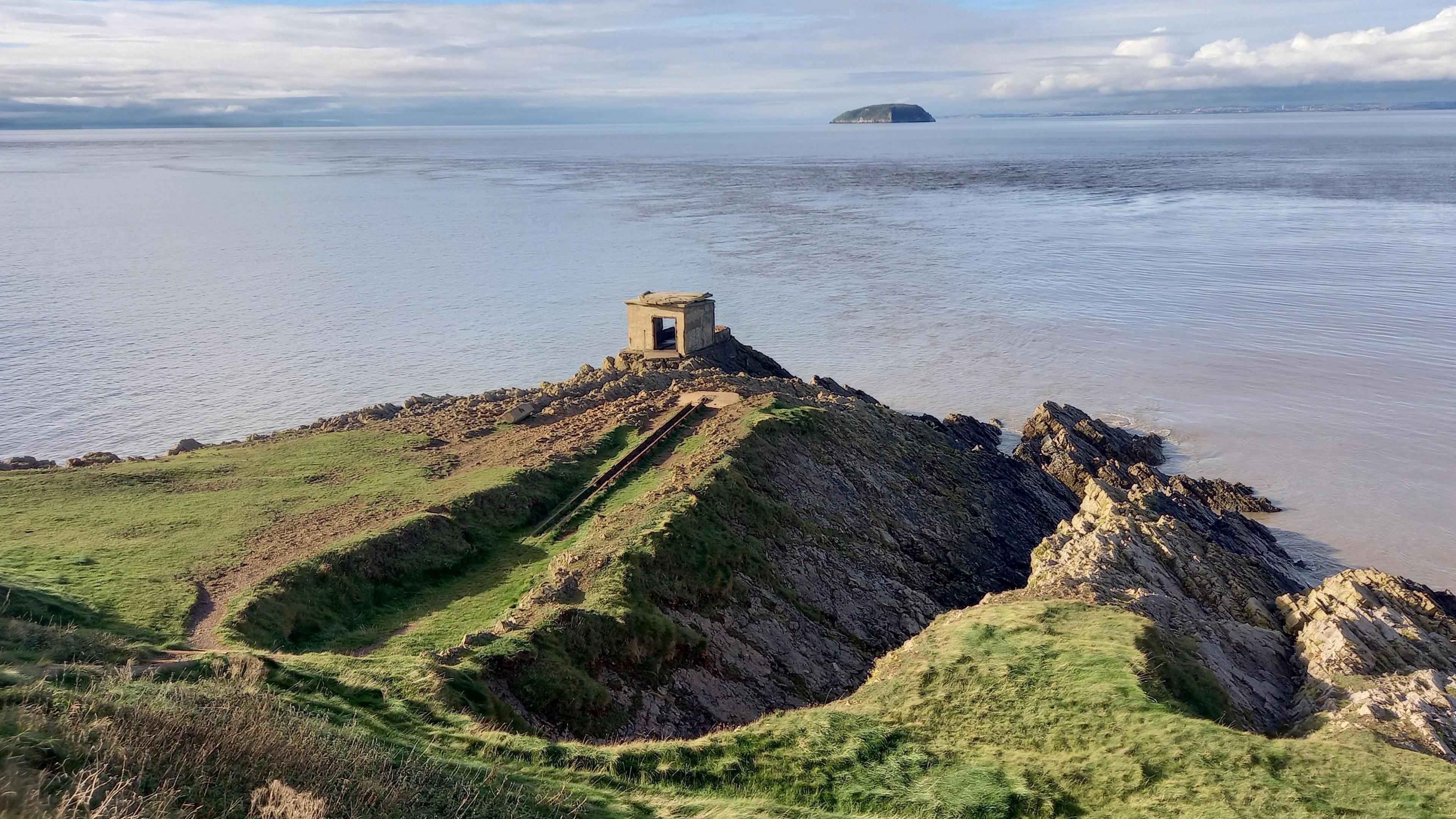 A rocky landscape of cliffs which are partially overgrown with grass and the sea in the background. There is a big rock poking out of the sea and a building is situated on the edge of the cliff