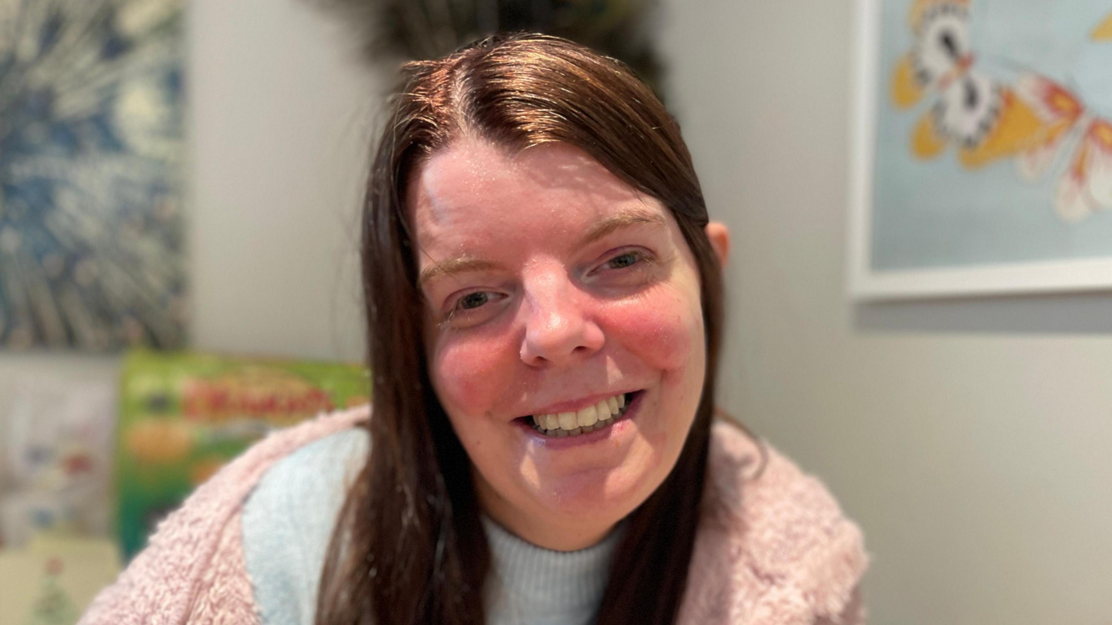 Kate is wearing a pink fluffy jumper and a grey sweater. Her long brown hair is worn down, with some of it tucked behind her ear. She's smiling at the camera.