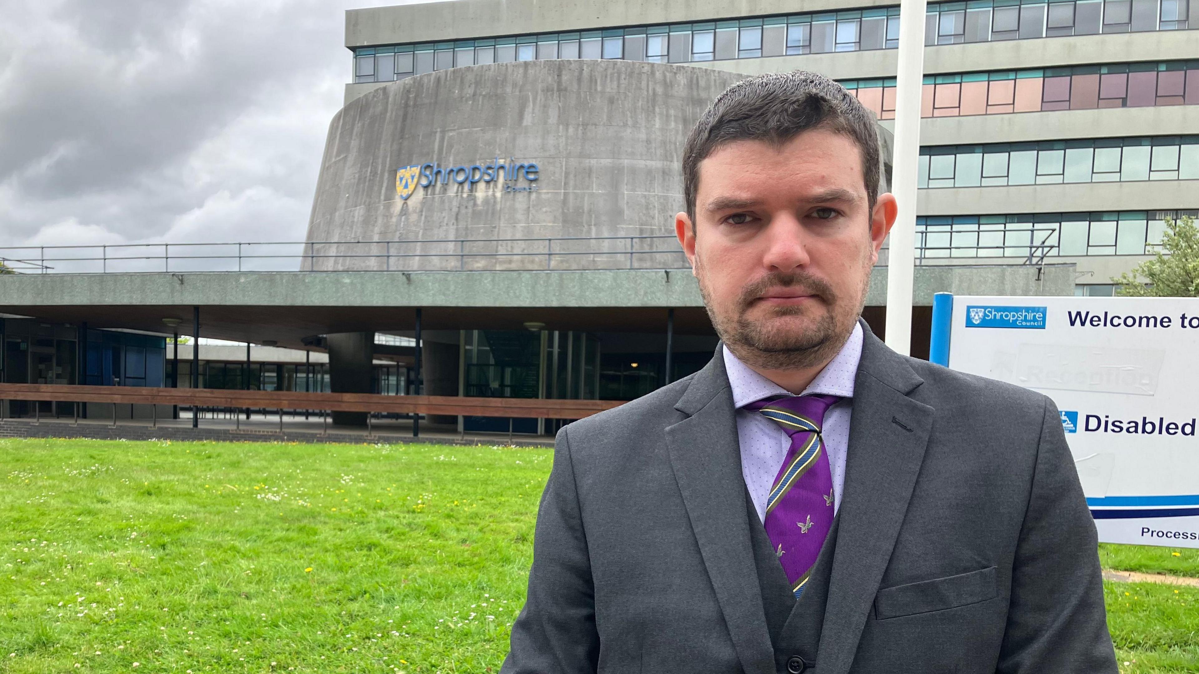 A man wearing a dark grey suit and purple shirt and tie  stands in front of a council building. In the middle distance is a building with a concrete cylinder on top with a sign that says Shropshire. Behind that is another building made up of concrete and glass