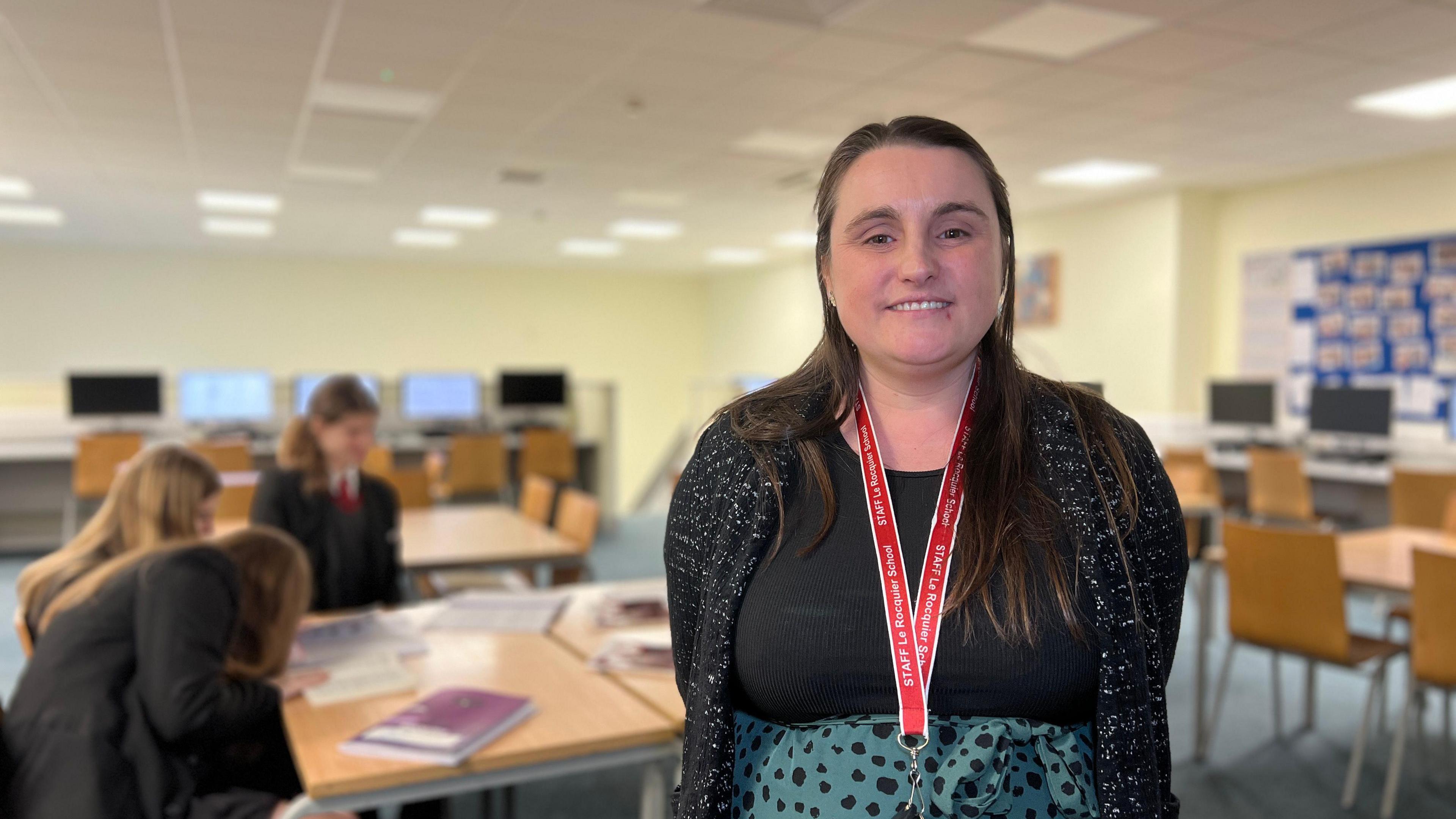 Gemma smiles at the camera with her hands behind her back. She has a brocade jacket over a black top with a spotted skirt. She has long dark hair which comes down to her chest and pupils are reading the book behind her.