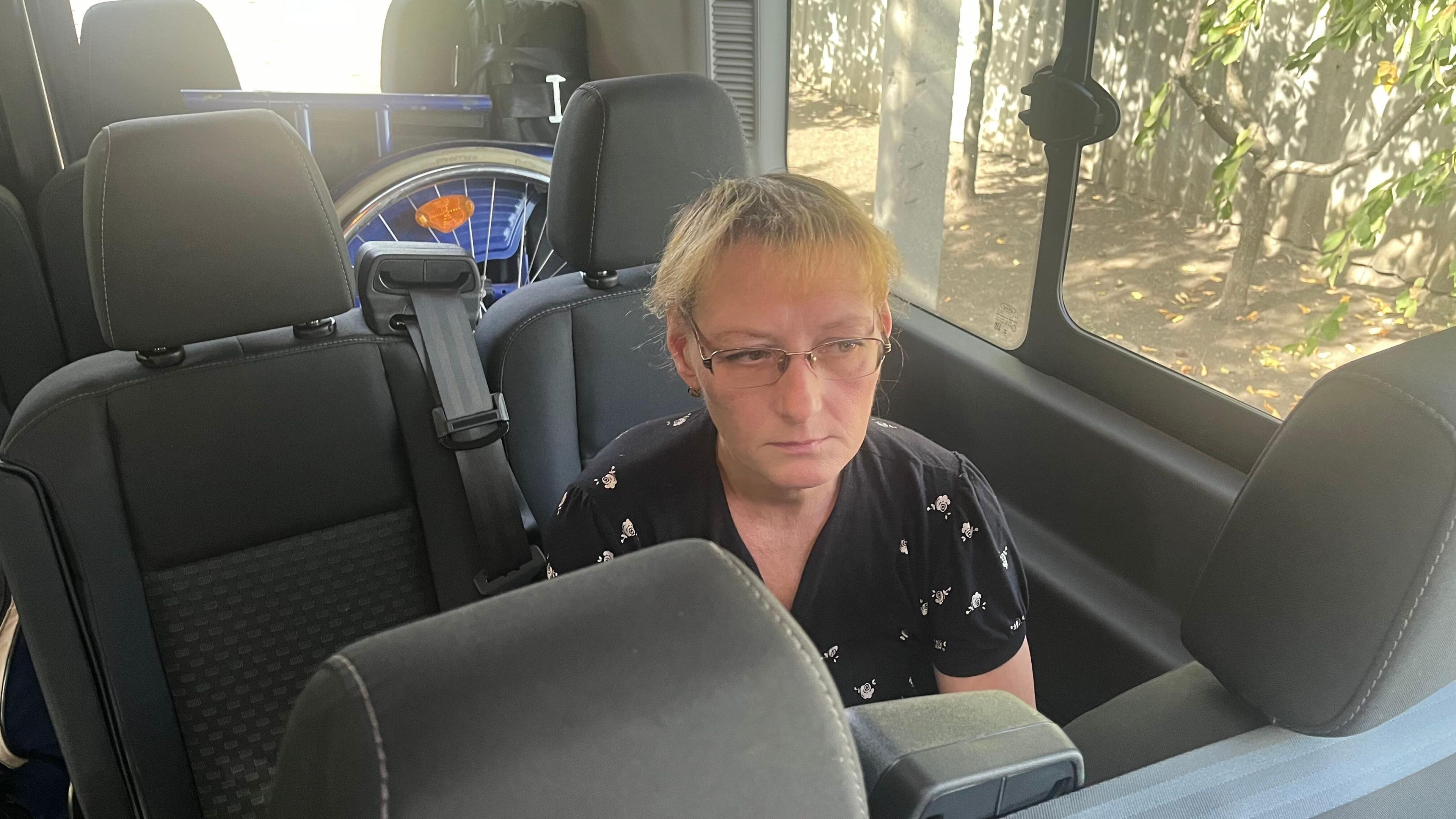 Viktoriia Vasylevska sitting in a seat of a vehicle - possibly a minibus. She is about 50 with blond hair and glasses, wearing a black blouse with a motif pattern.