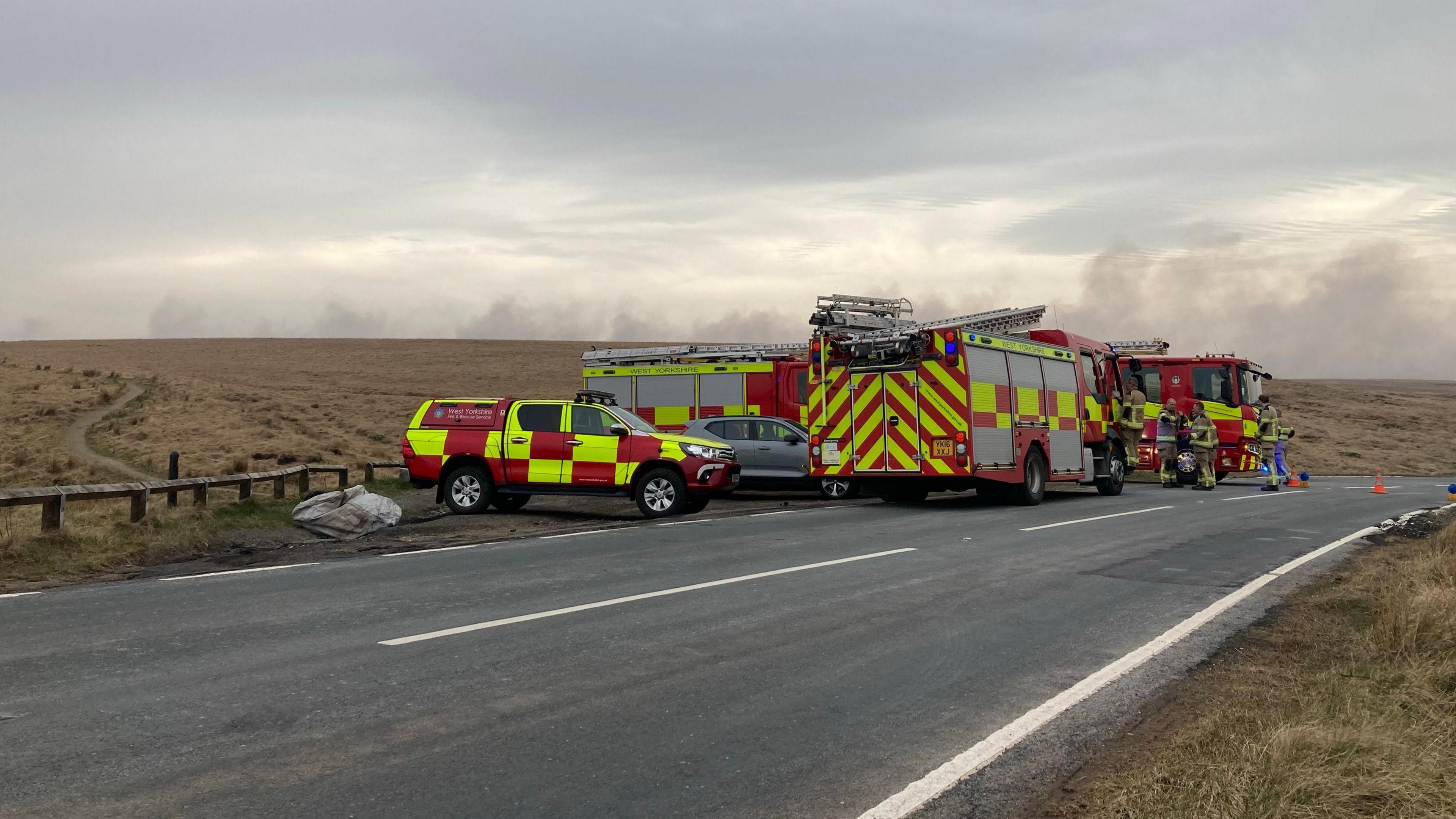 Fire vehicles parked on a road across a moor, with smoke rising in the background