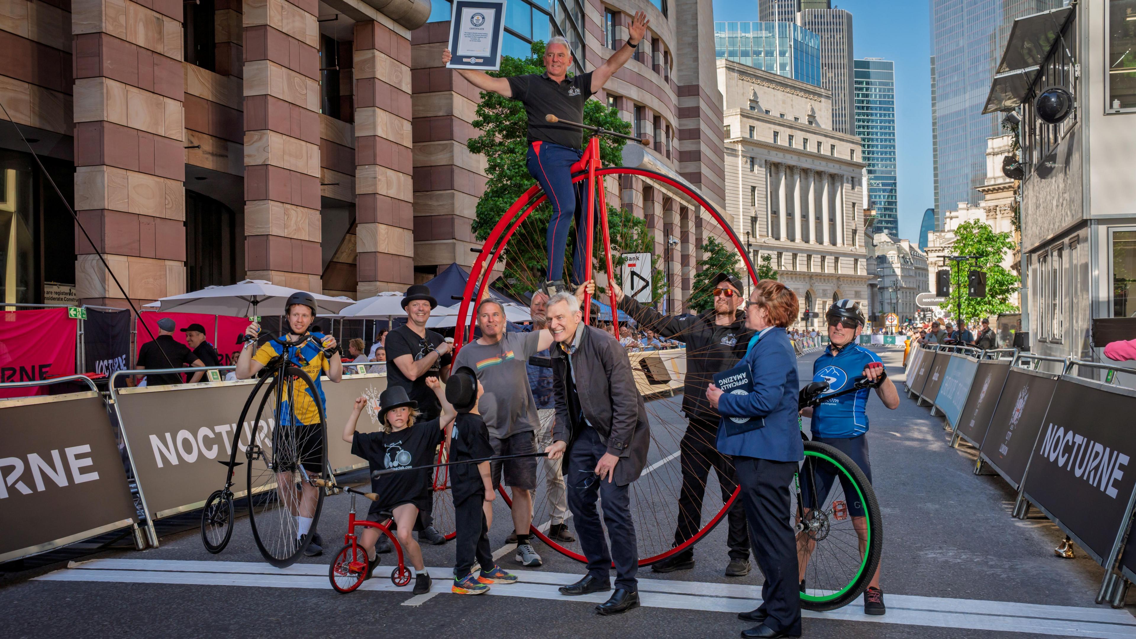 Giant penny farthing in red, tiny penny farthing also red, child cyclists - boy and girl - a juggling cyclist, a cyclist atop the massive bike, the Guinness World Records adjudicator. Everyone is smiling in the middle of a London street cordoned off for the event.