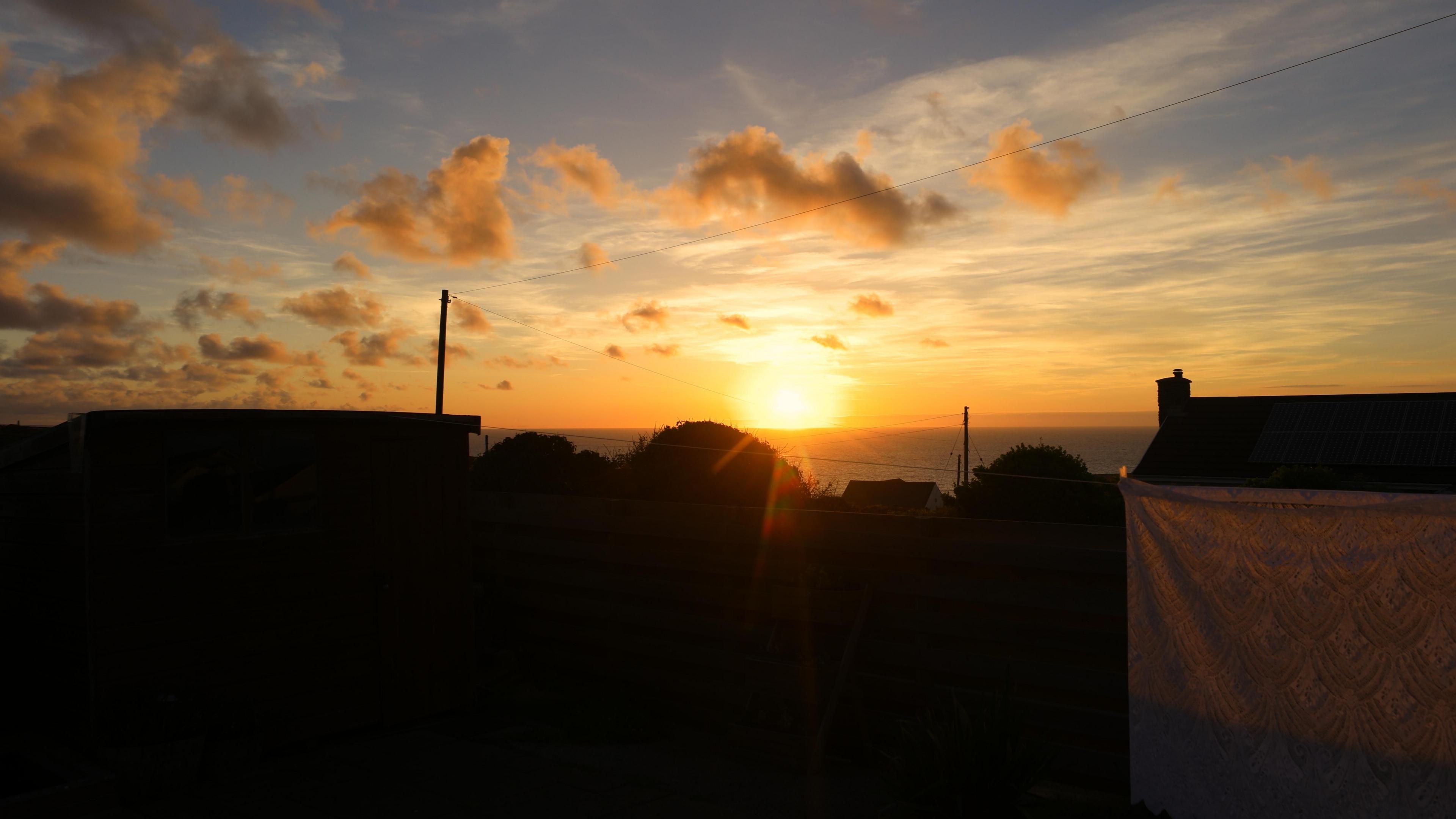 The sun sets over Porthtowan in Cornwall. The outline of a couple of houses and several trees can be seen. A bed sheet is handing on a washing line.