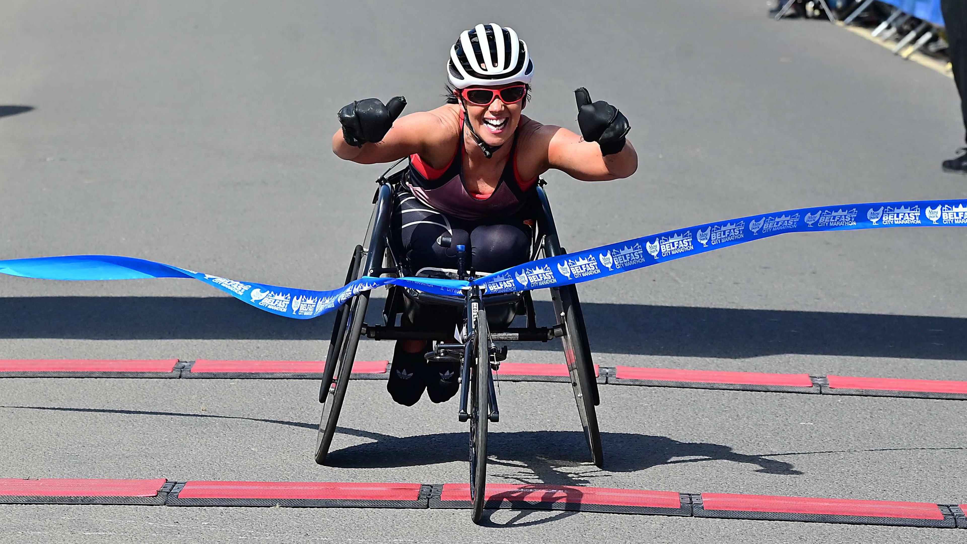 A lady in a wheelchair with a white helmet puts her thumbs up as she crosses through the blue ribbon. She is wearing red glasses and is wearing black gloves. She is wearing a vest with leggings.