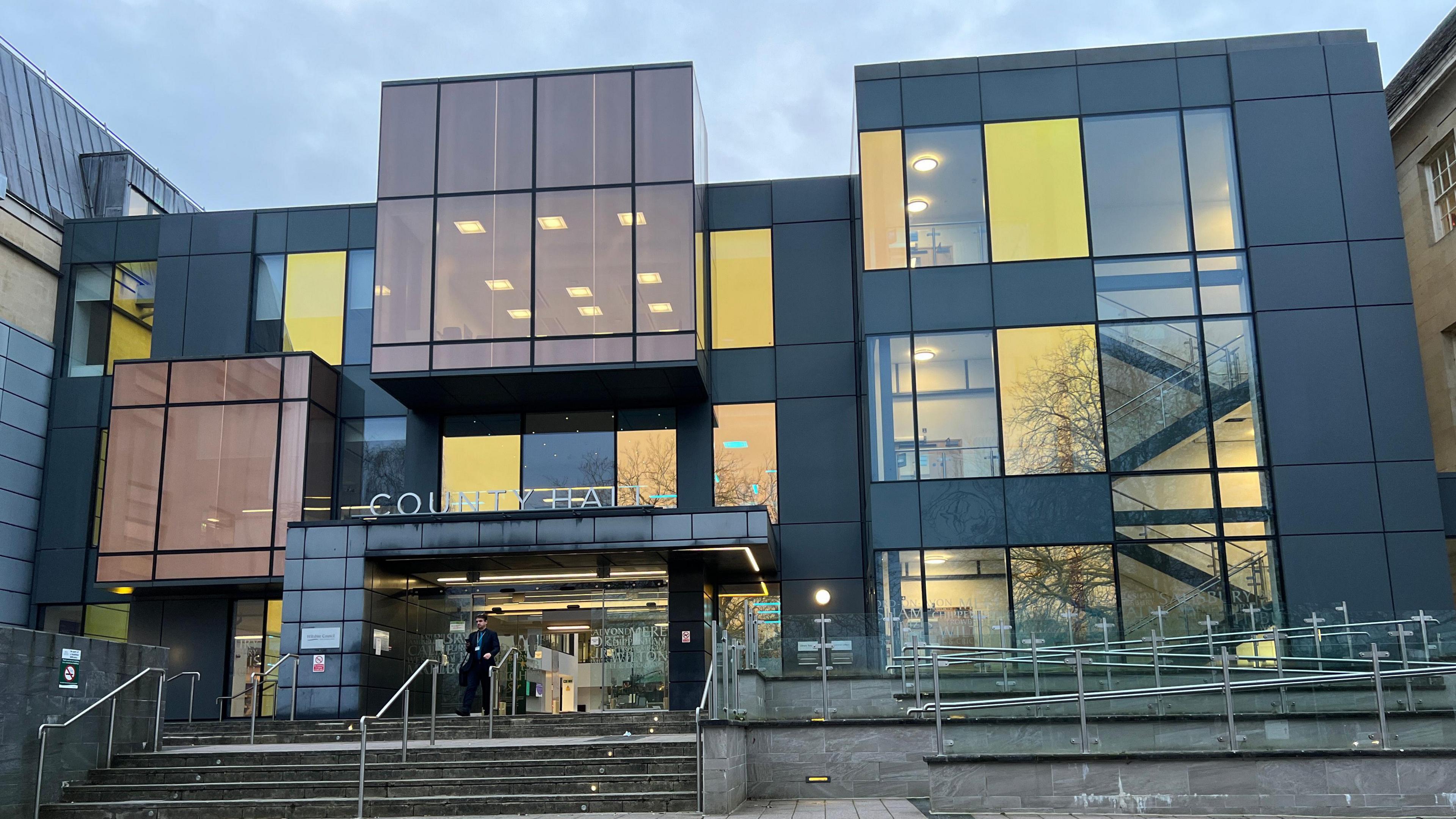 the front of a modernist building with multicoloured windows seemingly made up of geometric blocks with the sign "County Hall" and a staircase below