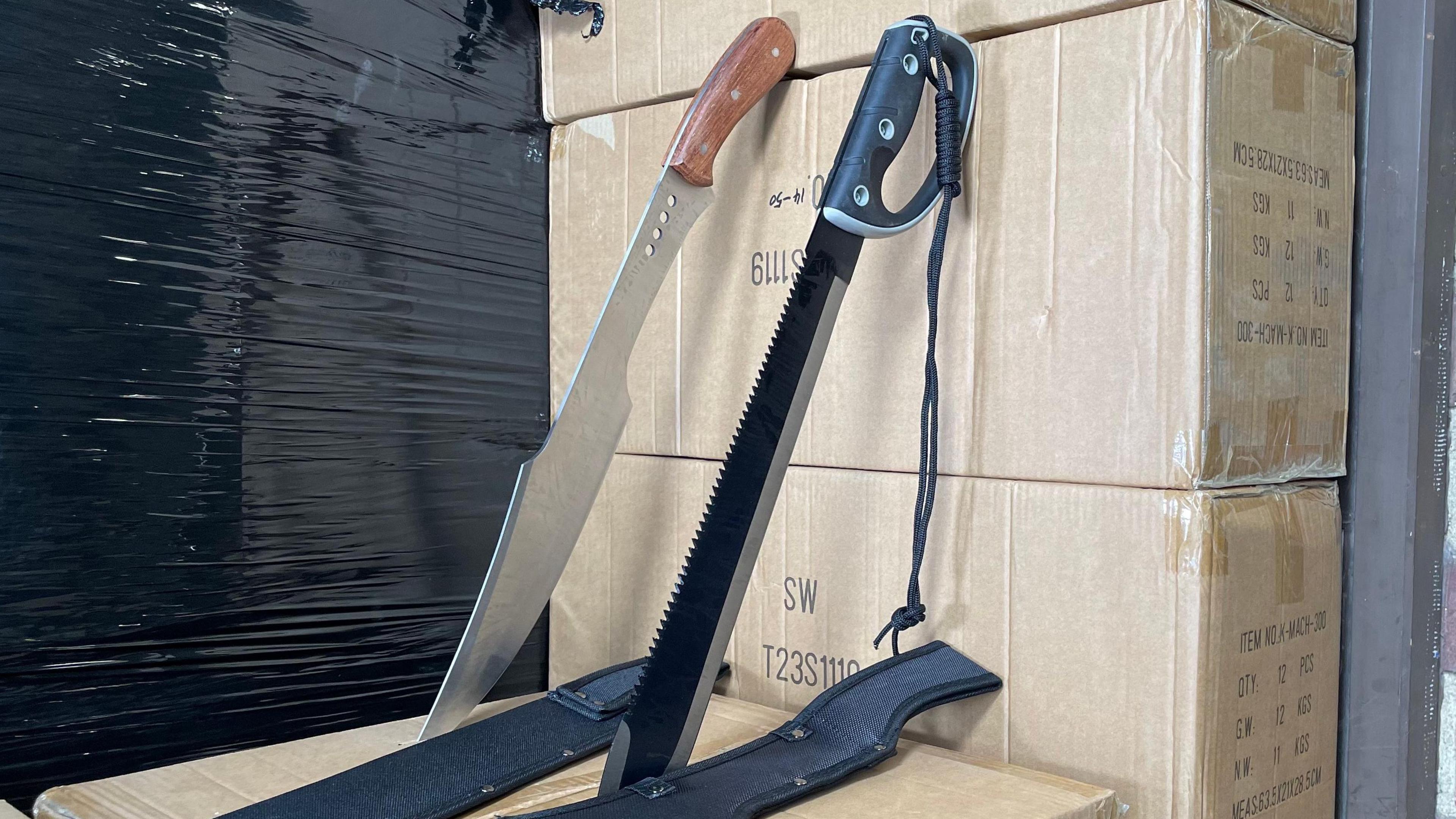 Two of the knives surrendered by Sporting Wholesale. They are both propped up, leaning against cardboard boxes, with the points digging into the top of another cardboard box. Both knives are around 18in (45cm) long. One is black, with a sharpened edge and a serrated edge. It has a black handle with a black rope hanging from the hilt. The other has a brown wooden handle, a blade that curves in places and four holes near the handle. 