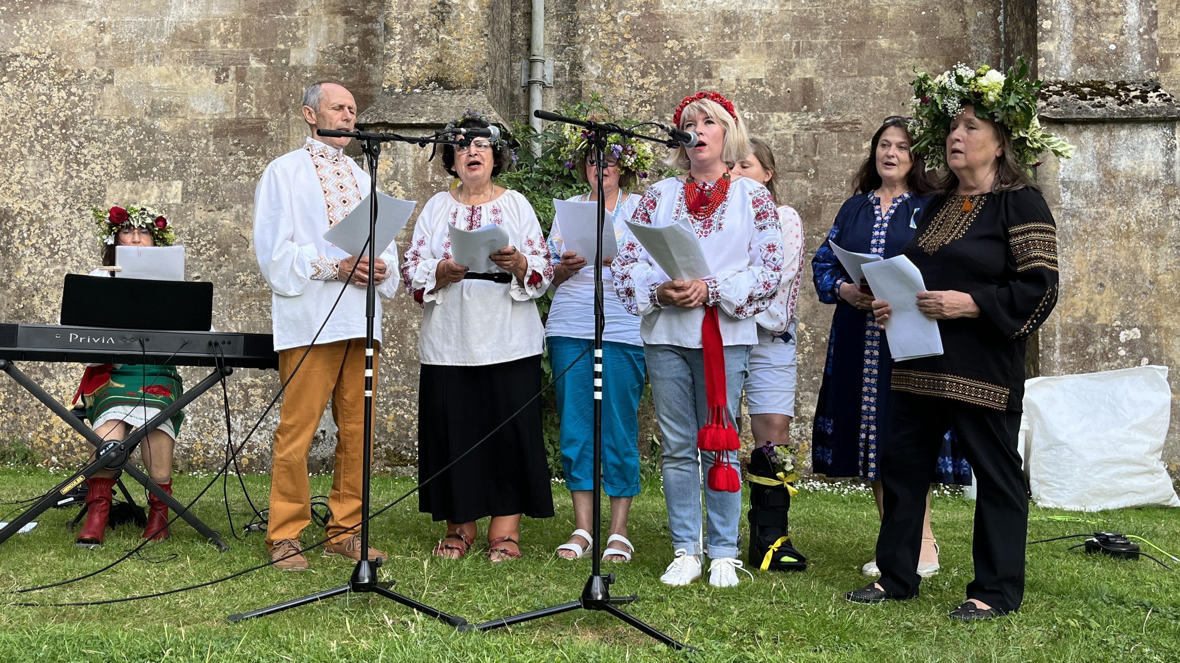 Malmesbury's Ukrainian choir wearing traditional clothing and singing into microphones in front of the Abbey wall.