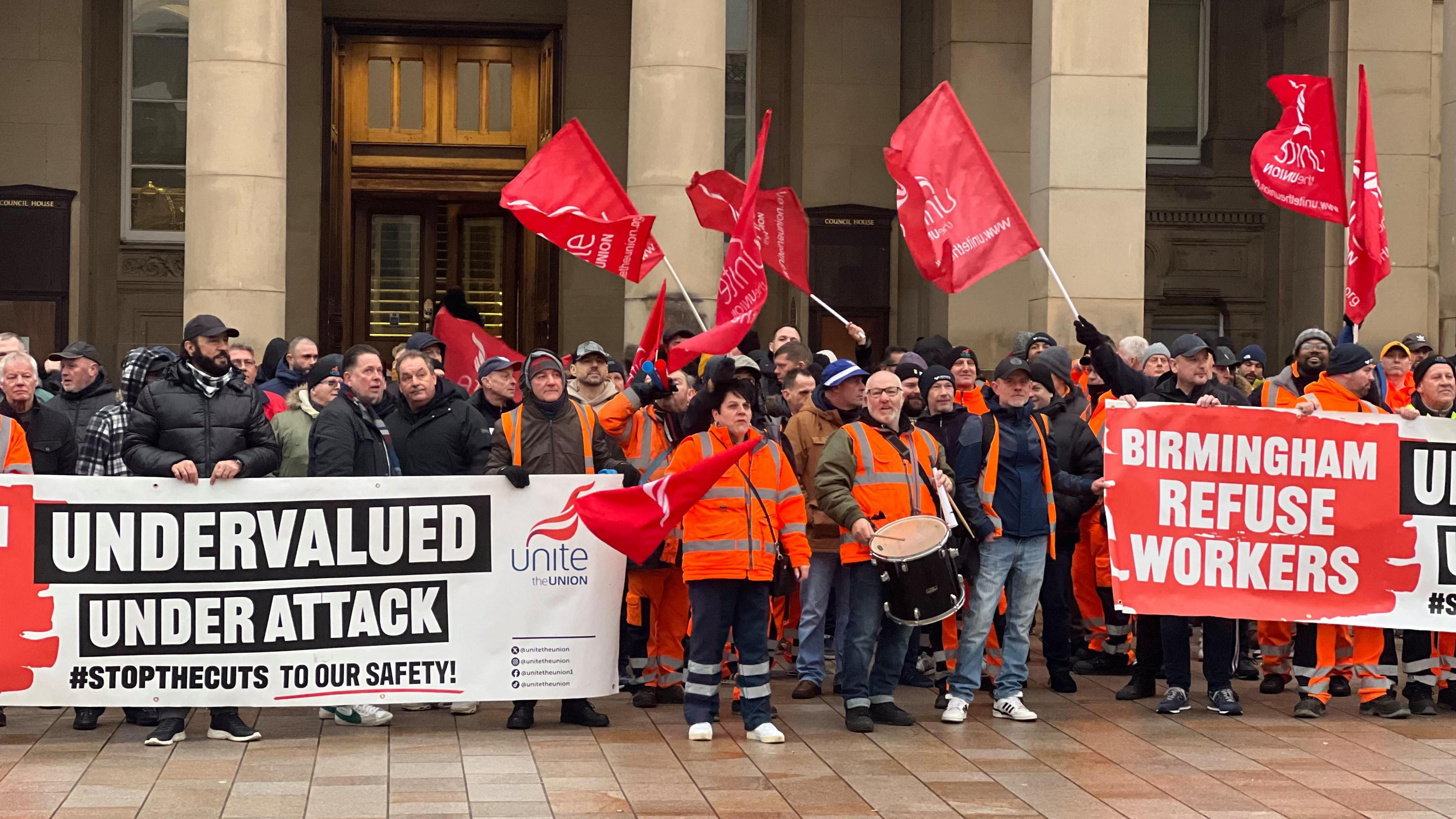 A group of people, some in hi vis clothing, stand outside the columns of a building. They are flying red flags and holding signs reading: "Undervalued. Under attack. hashtag stop the cuts to our safety".