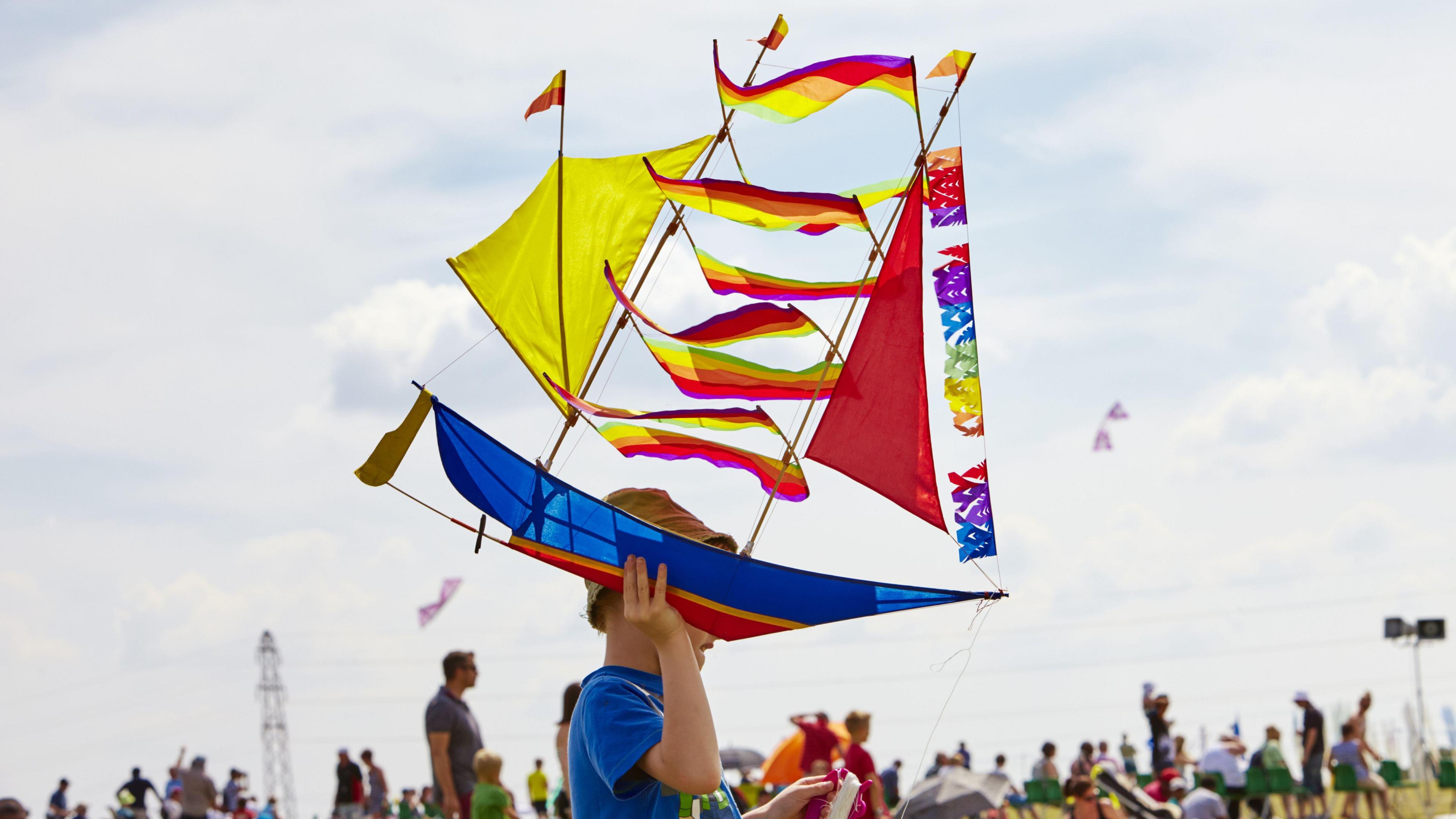 A child, wearing a hat, with a blue top on, holding a colourful kite, in the shape of a boat