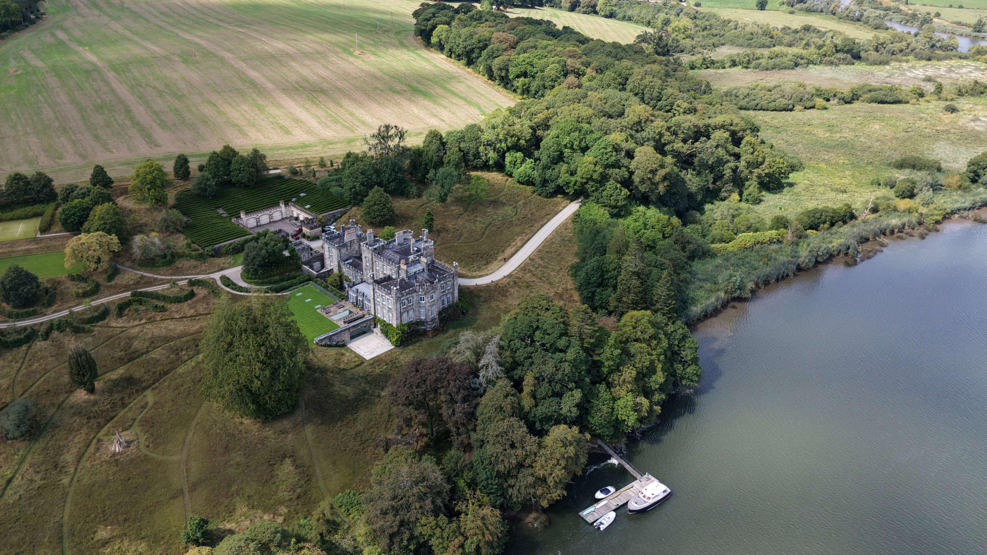 An aerial view of a castle, surrounded by trees and fields. It is overlooking a river. There is a boat on the river.