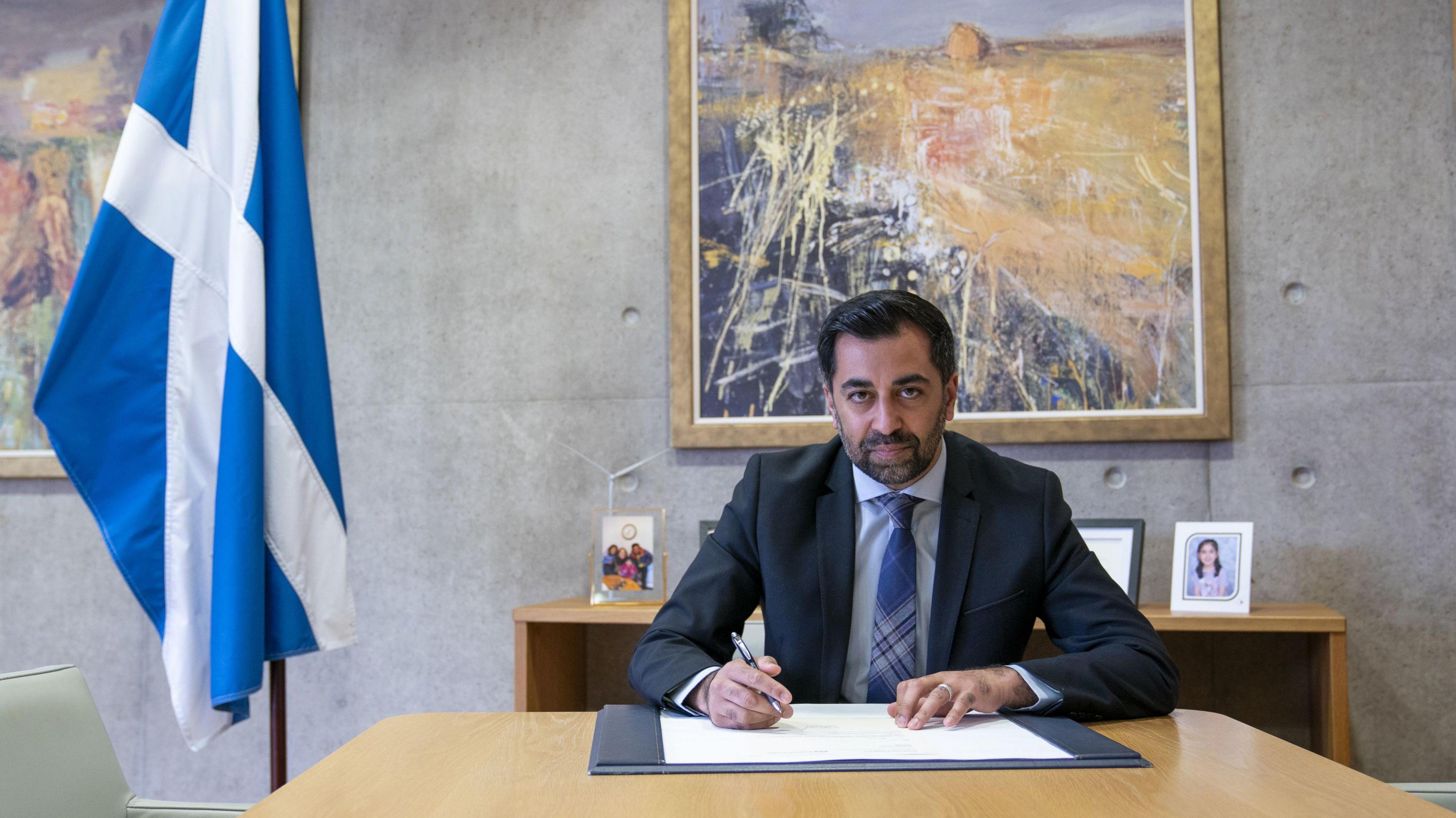 Humza Yousaf is sitting at a bare, pale blonde wooden table in his former office in Holyrood. He is about to sign a paper. He is dressed in a blue suit, blue shirt, blue tartan tie. There is a furled saltire on the left. Family photos can be seen in the background.