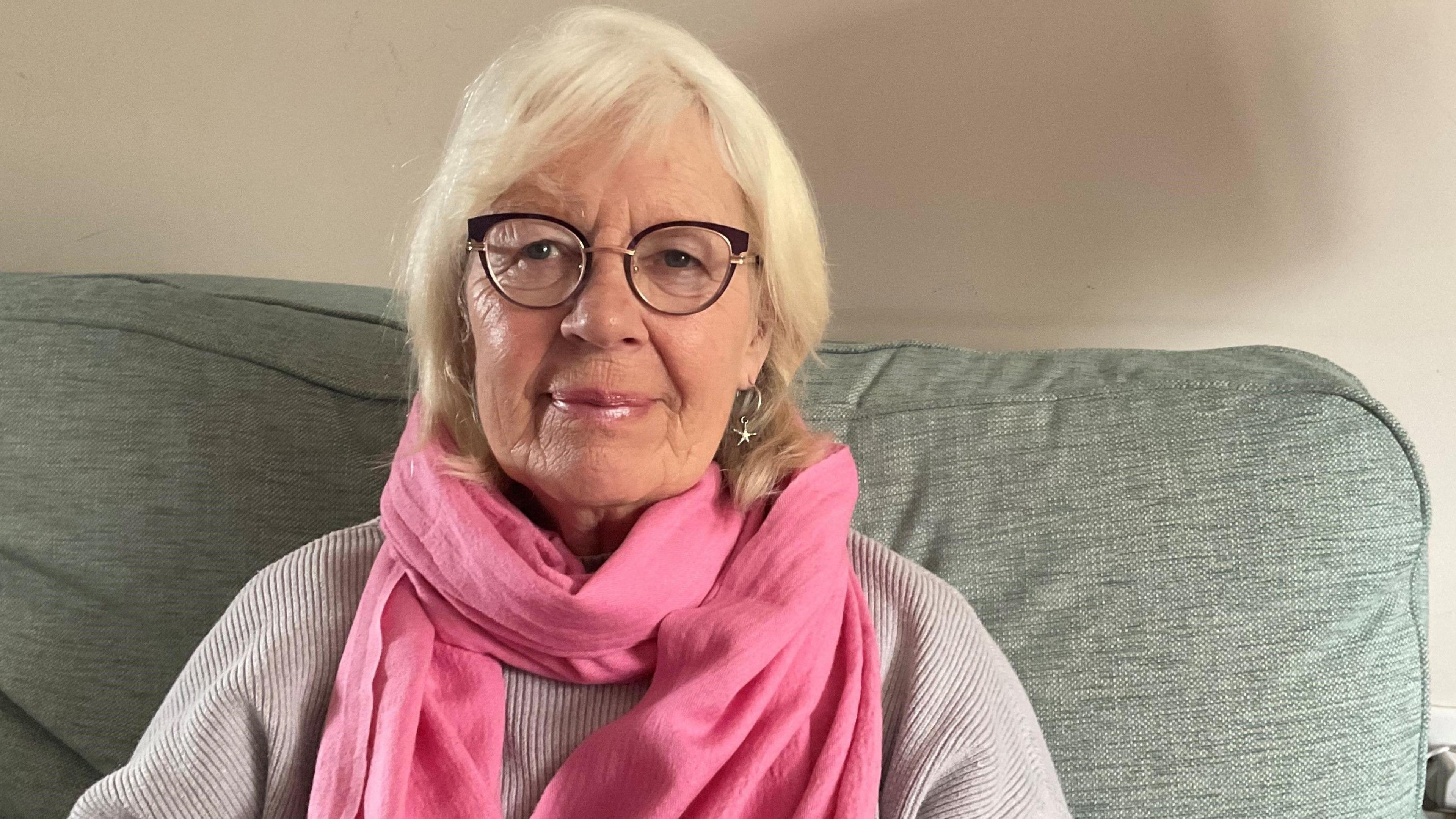 Karen Langford smiles as she sits on a grey sofa wearing a pink scarf and jumper