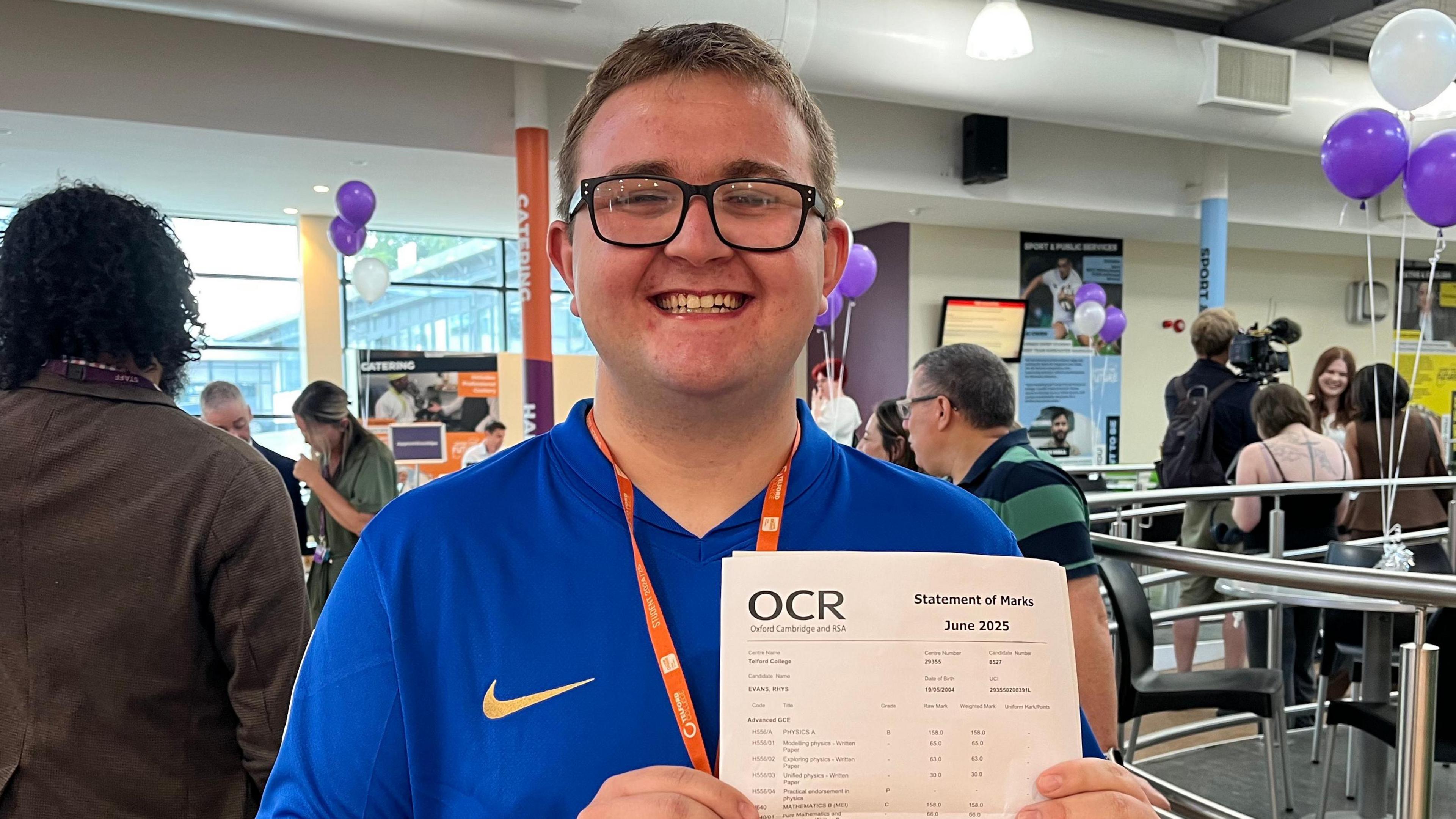 Rhys holding up his results - a white piece of paper with little detail visible, but the letters 'OCR' are written in bold in the top left hand corner. Rhys is wearing a blue sports shirt, an orange college lanyard, and he's stood in a school atrium which has white walls and floor-to-ceiling windows. There are a few bunches of purple and white balloons dotted around the room. 
