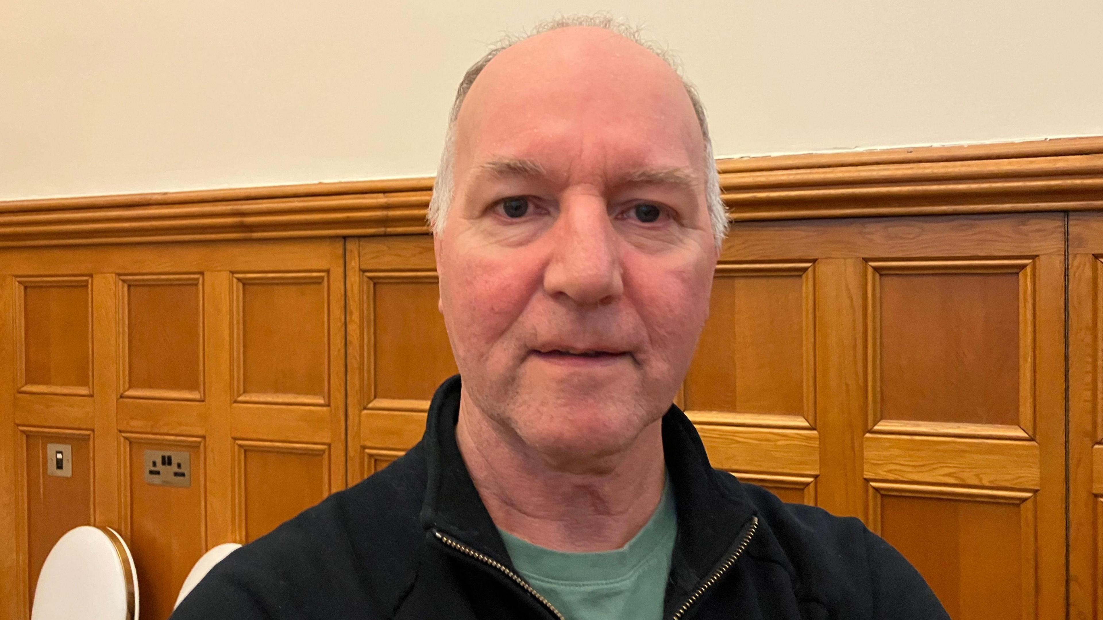Tony Doherty is wearing a green shirt under a black jumper. He has patchy grey head hair. Behind him the wall is paneled with wood. There are two cream chairs sitting. The wall is painted an off-white shade.