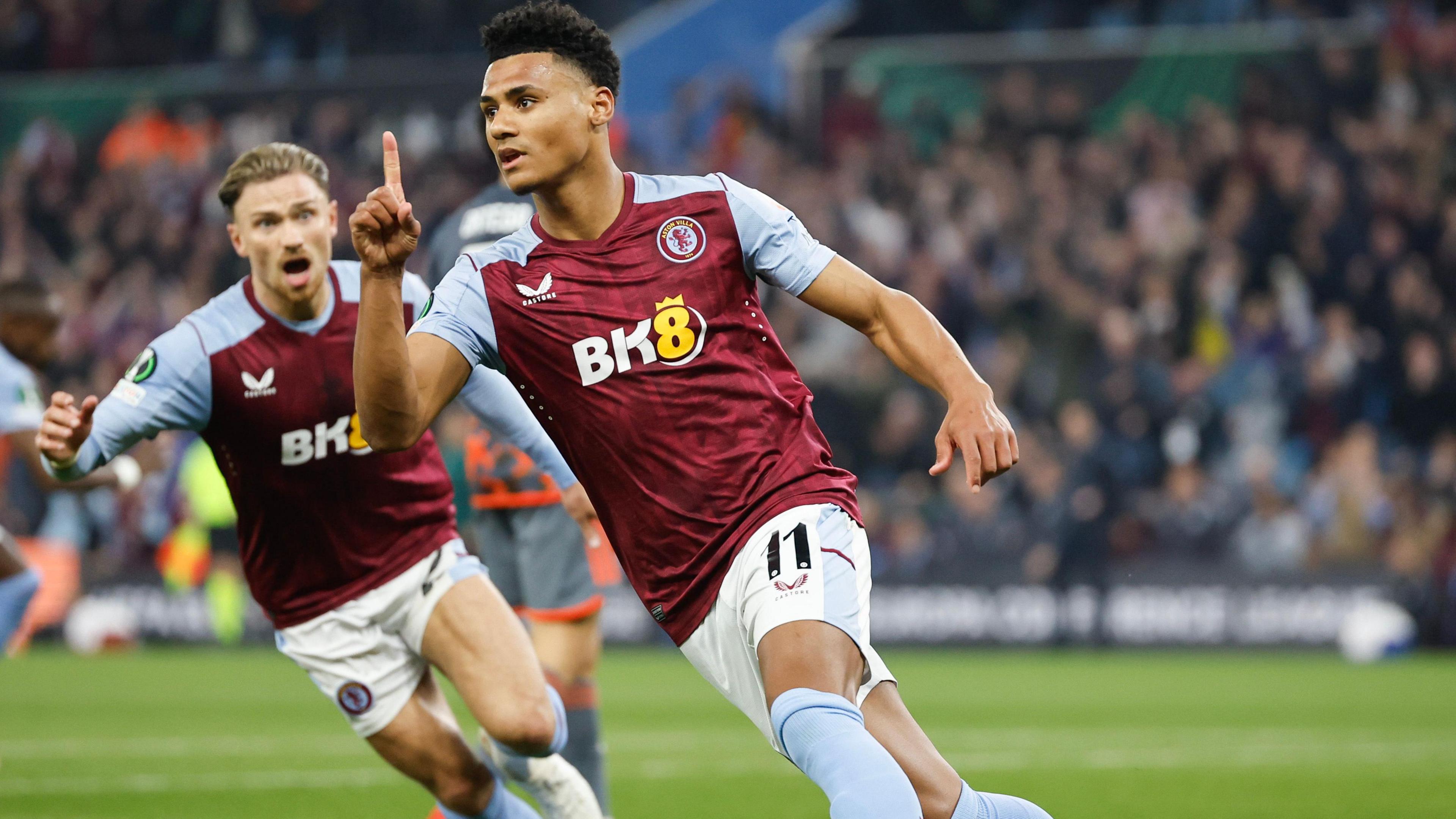 Ollie Watkins turns to celebrate with his right hand in the air, displaying his index finger, with Matty Cash celebrating behind him.