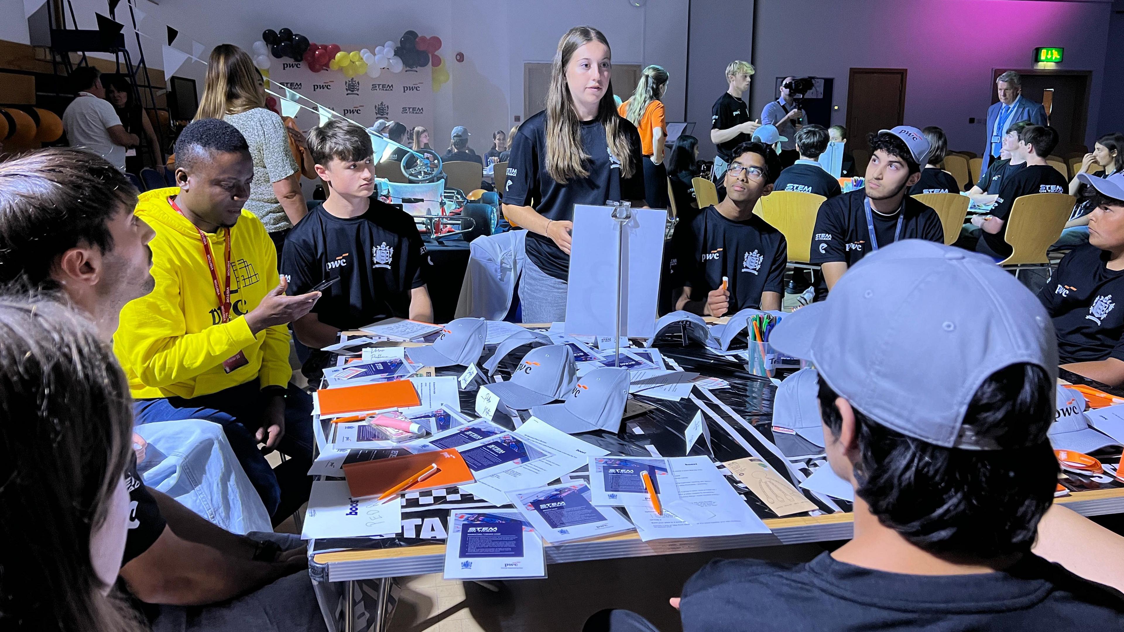 A group of people gathered around a table covered with papers and notebooks, engaged in conversation and collaborative activities. The setting appears to be a workshop or group event, with additional people and balloons visible in the background.