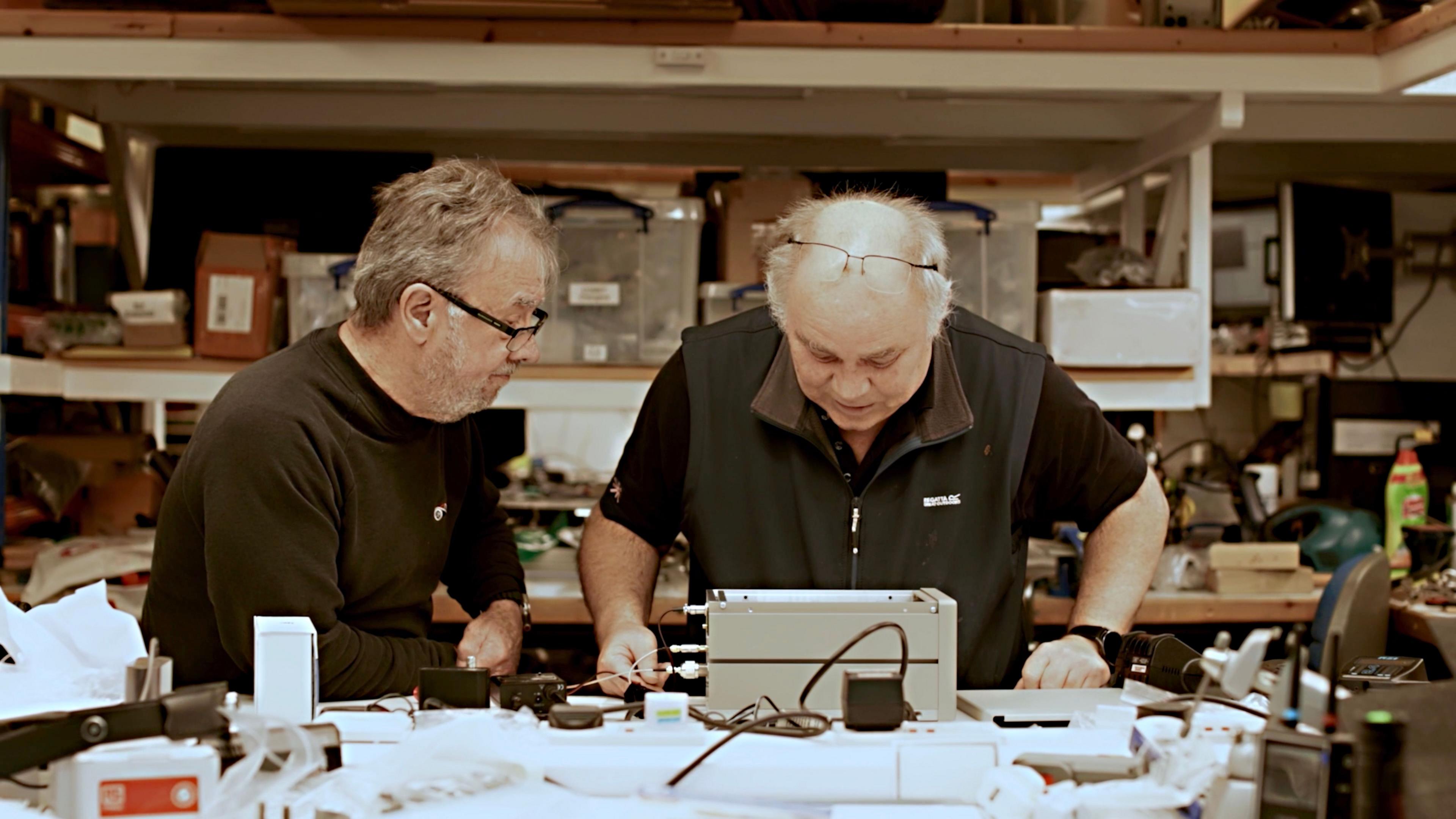 John Little and Greg Willaims working on The Thing, in a workshops with materials all around them, including boxes behind them and electronic equipment. John, left has a black top on and glasses, and Greg, glasses on his head and a black top and gilet. They are both looking at a piece of equipment.