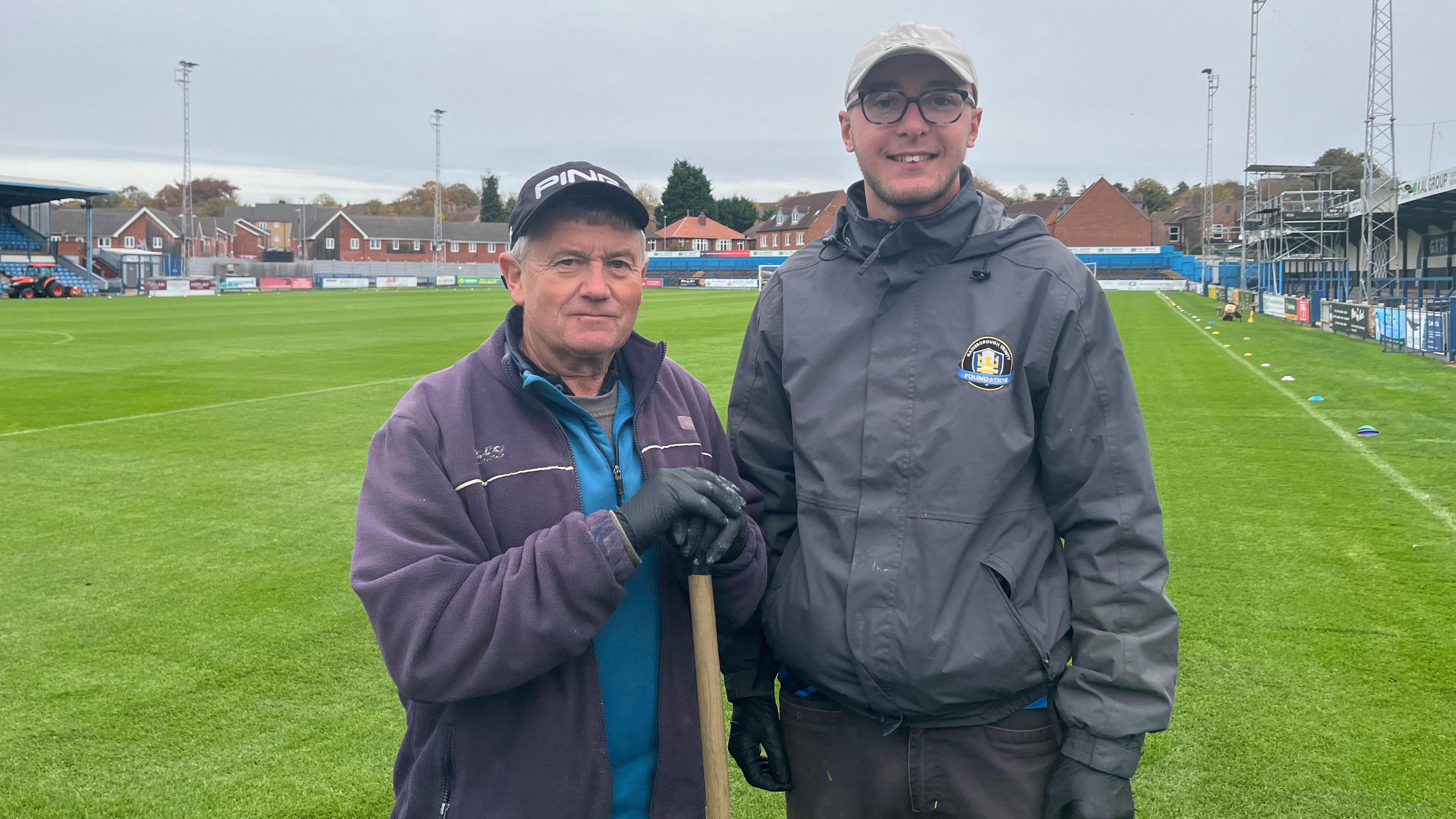 An older man wearing a black Ping cap and a black and blue fleece is wearing gloves with his hand resting on a wooden pole. Next to him is a taller and younger man in a cap and a black jacket with a football logo on it. Behind them is an empty football pitch.