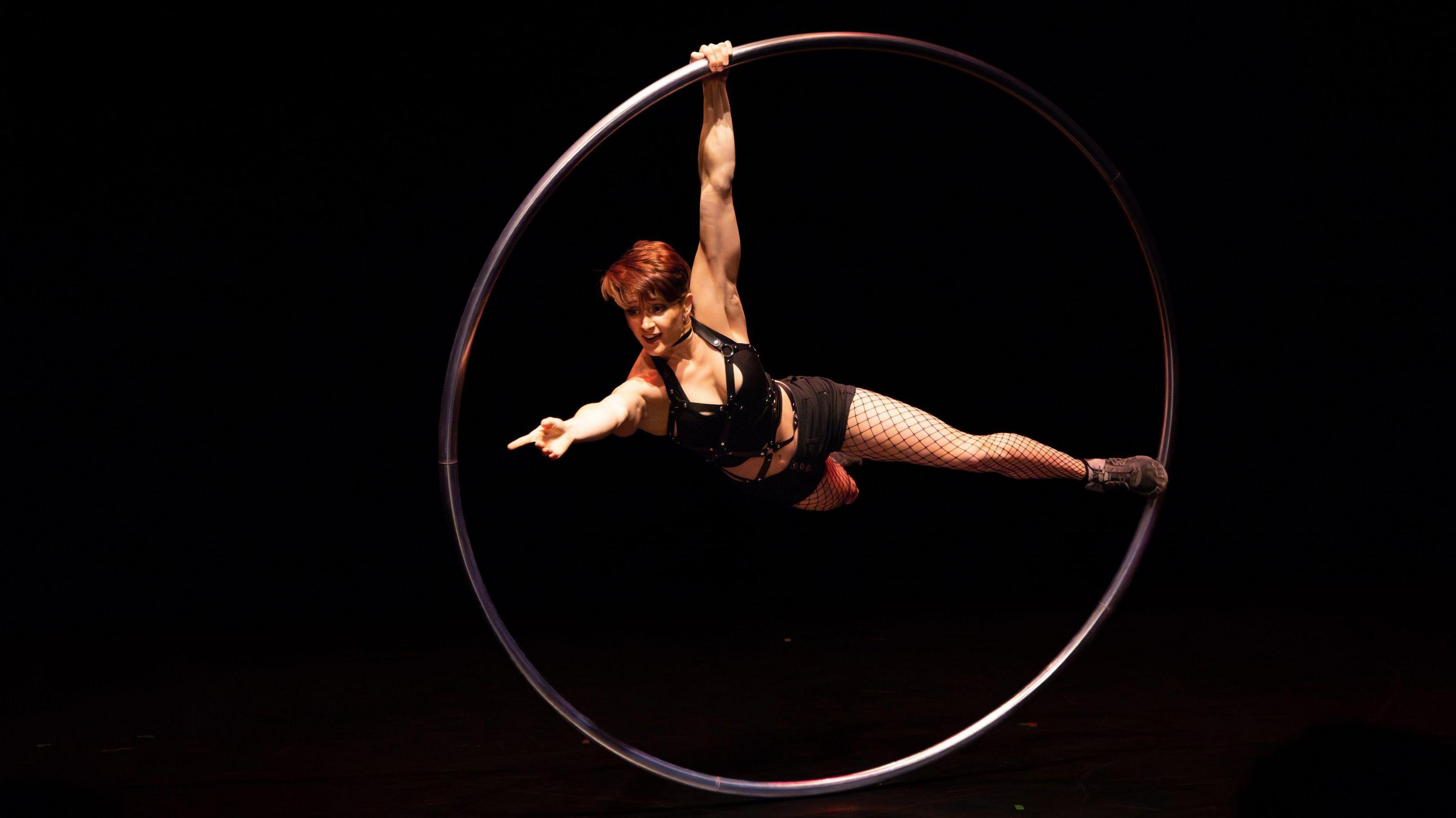 A woman in a black outfit swings inside a giant metal circle as part of the Decadent Rabbit show that is being performed by Circomedia in Bristol. She has short cropped hair and is pointing into the distance with one arm