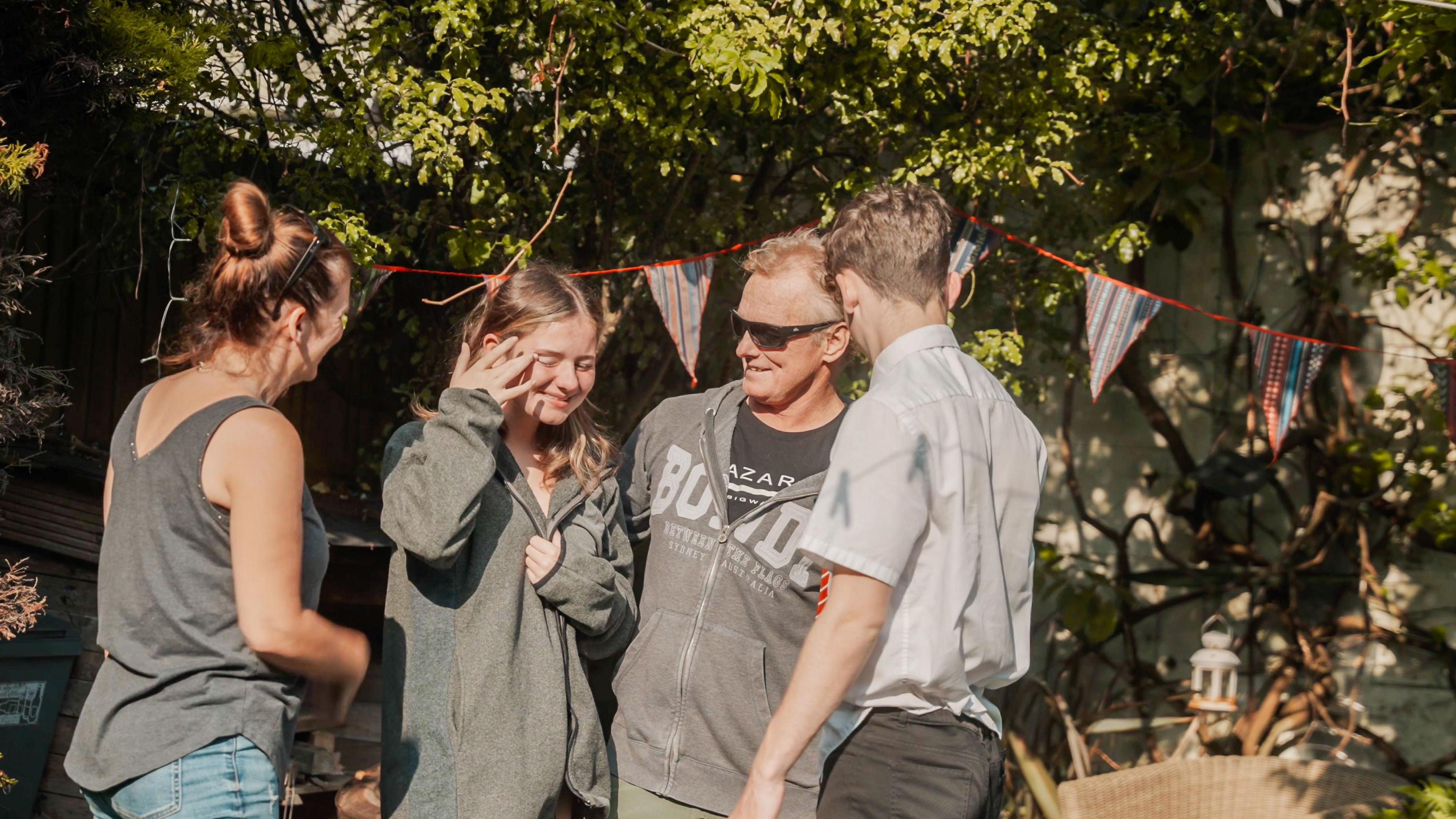 Mark returning home after his second stem cell transplant. He is in black sunglasses and a grey hoodie and is stood next to his daughter, also in grey hoodie with brown hair, his wife, who is in a grey vest top and is facing the wrong way and his son, who is in a light grey shirt and also has his back to camera