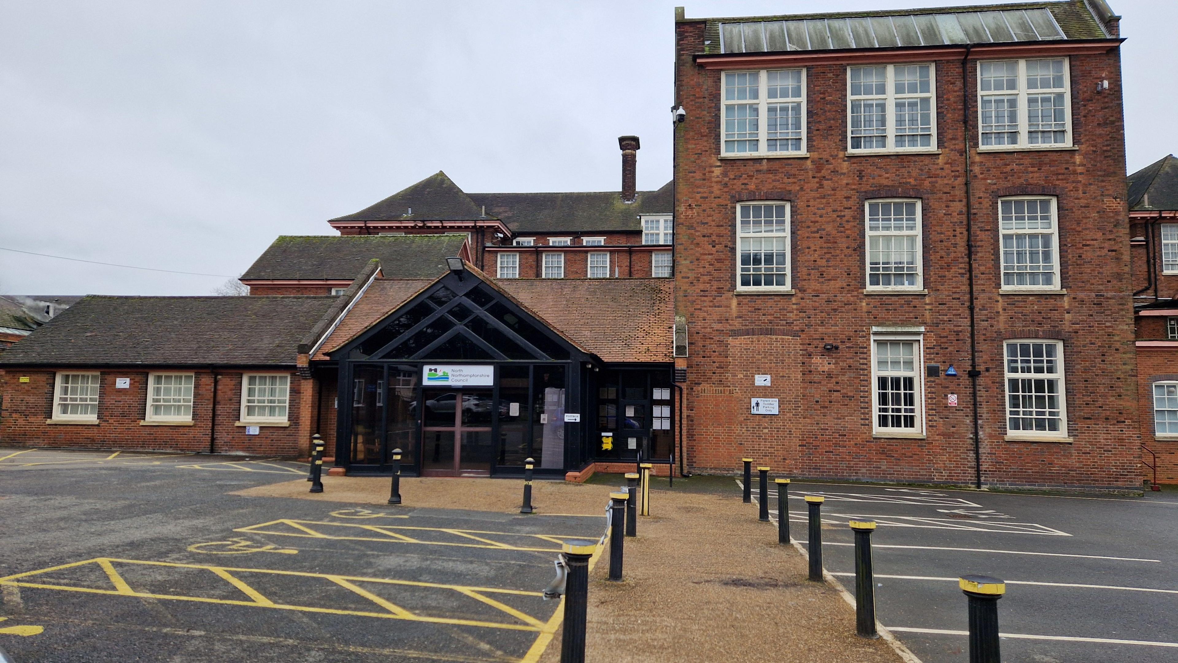 A brick building with three storeys on the right one storey on the left. There is a car park and a path in front.