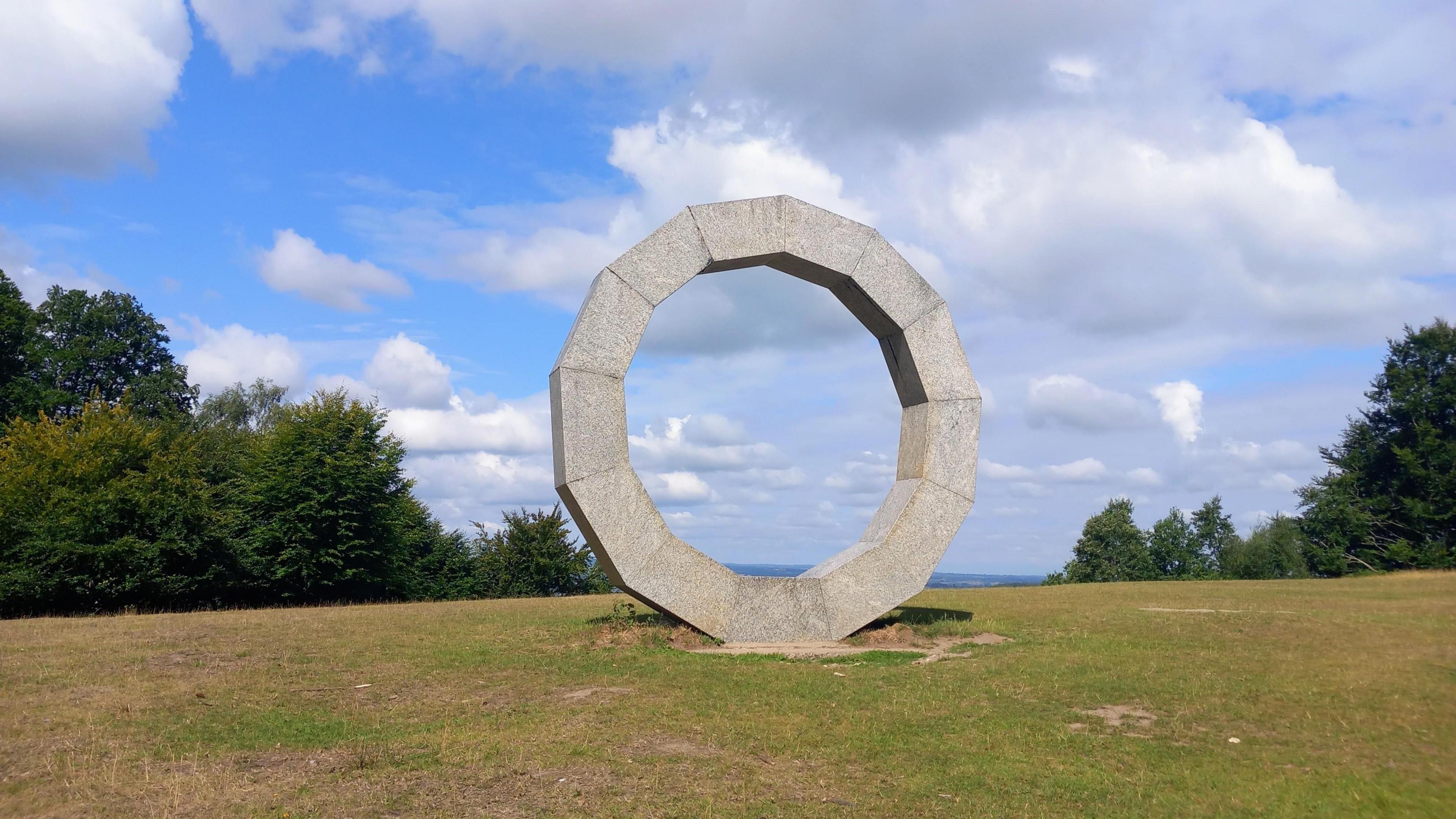 A ring-shaped structure made of granite standing on a hillside with trees on either side and an obscured view across a valley in the background.