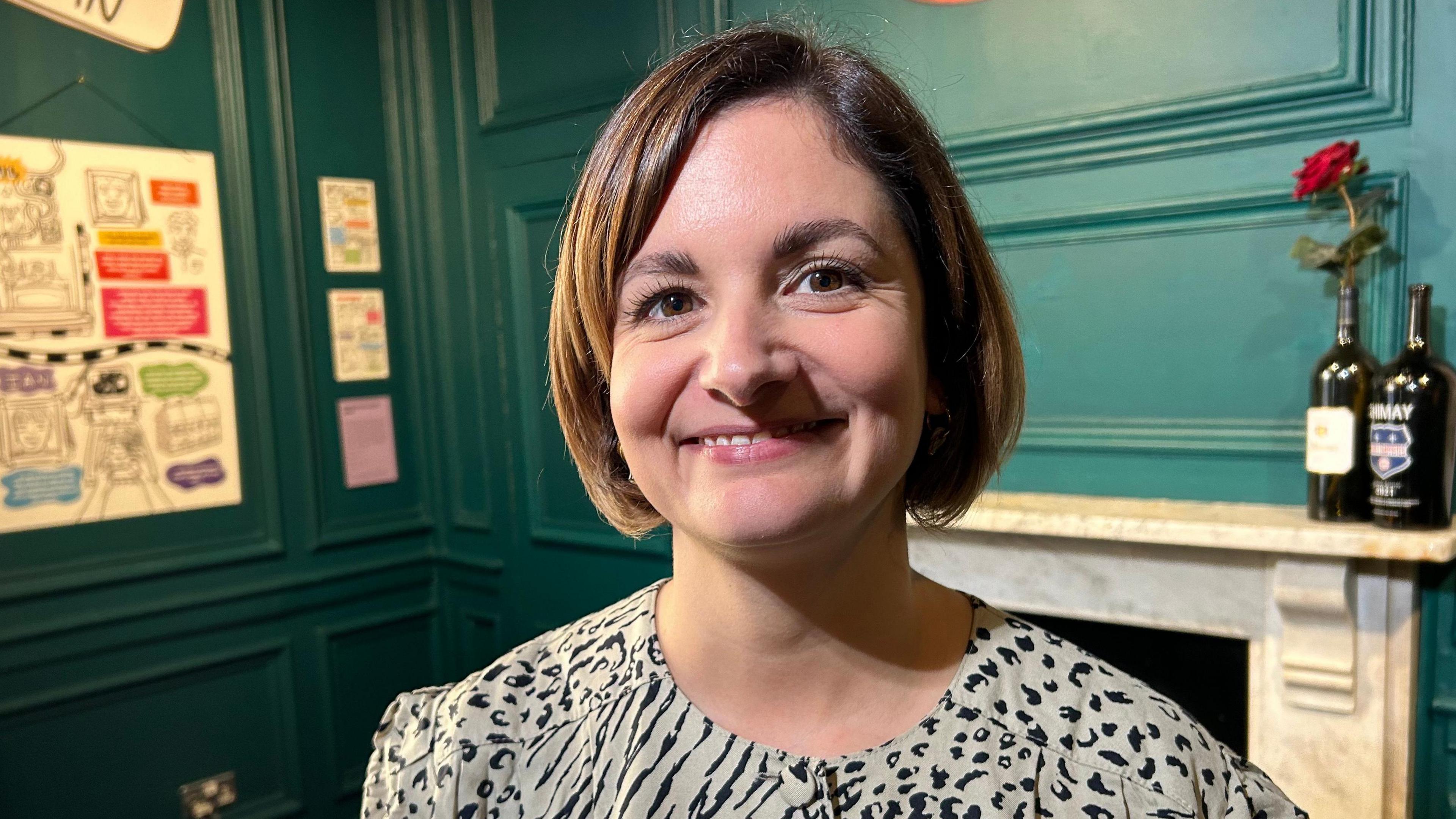 A woman with short brown hair and brown eyes. She is smiling widely at the camera and is wearing a black and white shirt in an animal print style.